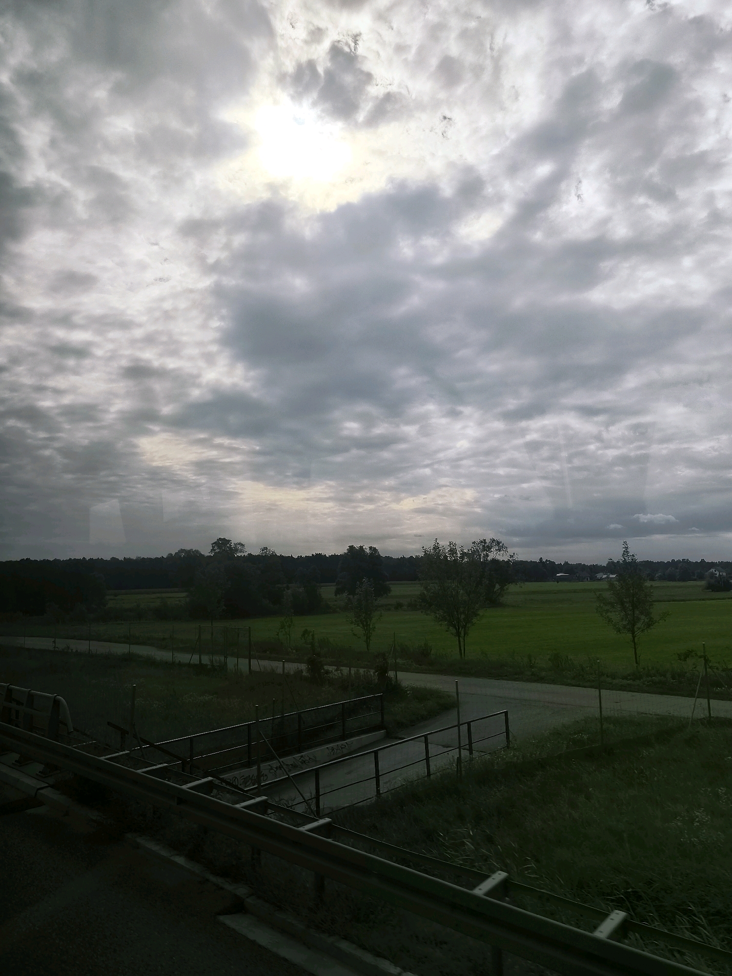 The image shows a landscape view with a cloudy sky. In the foreground, there is a metal railing that appears to be part of a barrier or bridge. Beyond the railing, there is a road and a grassy area. In the middle ground, there is a field of green grass bordered by trees. The background consists of a dense line of trees and a faint suggestion of buildings or structures. The sky is overcast with patches of sunlight breaking through the clouds. The scene appears to be taken from a moving vehicle, likely a train or car, given the perspective and the visible railing. There are no people or animals visible in the image. The location is not specifically identifiable as being in Królewiec, Poland, as there are no distinctive landmarks or signs.