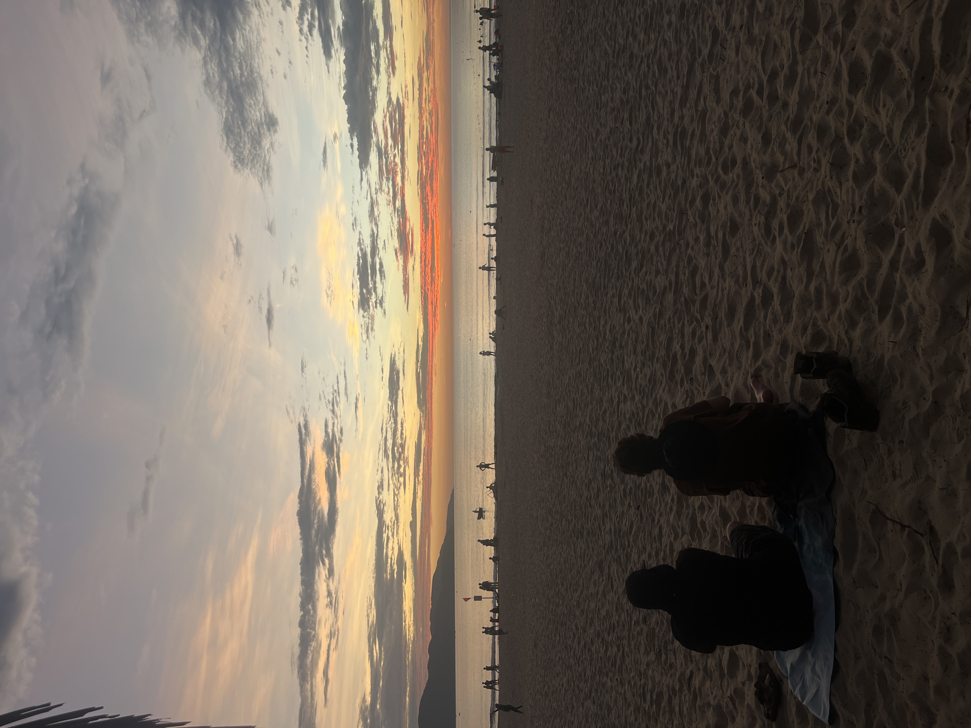 The image captures a beautiful sunset scene on a beach, likely in Đà Nẵng, Vietnam. The horizon line, stretching across the middle of the frame, separates the sky from the ocean and sandy shore.
In the immediate foreground, two individuals are silhouetted, seated on the textured sand, facing towards the water and the setting sun. One appears to be sitting on a light-colored cloth or towel. Their relaxed postures suggest they are observing the evening panorama.
The sky is a prominent feature, showcasing a dramatic sunset. Hues of fiery orange, yellow, and soft purple blend across the horizon, contrasting with scattered dark grey clouds that add depth and mood to the upper sky.
Along the distant shoreline, numerous small figures are visible as dark silhouettes against the bright water and sky. These people are scattered across the beach and some appear to be in the shallow ocean, enjoying the twilight. A small red flag or marker can be seen near the water's edge in the mid-ground.
In the far distance, a range of mountains rises, silhouetted against the colorful horizon. The ocean surface reflects the fading light, appearing calm and extending towards the vibrant sky. The sandy beach in the foreground shows subtle undulations and footprints. The overall atmosphere conveyed is peaceful and contemplative, capturing the serene beauty of dusk at the coast. No visible text is present.