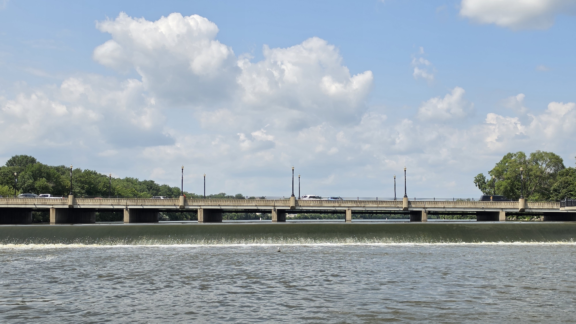 An eye-level, medium shot shows a concrete bridge over a dam with water flowing over it in Elgin, United States of America. The bridge, which spans the horizontal width of the frame, has a concrete railing and is supported by several concrete piers. Lampposts are visible along the top of the bridge, and cars are driving on it. The water in the foreground is choppy and has white foam where it flows over the dam. Trees and green foliage line the far bank of the river behind the bridge, and a partly cloudy blue sky fills the upper portion of the frame. A single bird is visible in the water in the foreground, and there are no people in the image.
