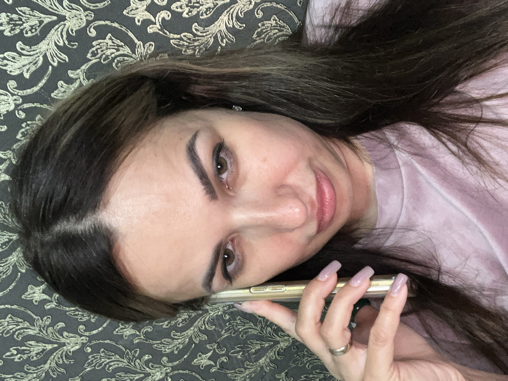 A woman is taking a selfie with her phone in hand, her gaze directed slightly downwards, with a neutral expression. She is indoors, with a patterned wallpaper visible in the background. The woman has long, dark brown hair and is wearing a pink t-shirt. She has a ring on her left ring finger. The lighting suggests an indoor environment, likely natural light from a window or artificial room lighting. The image captures a close-up of her face and upper body.