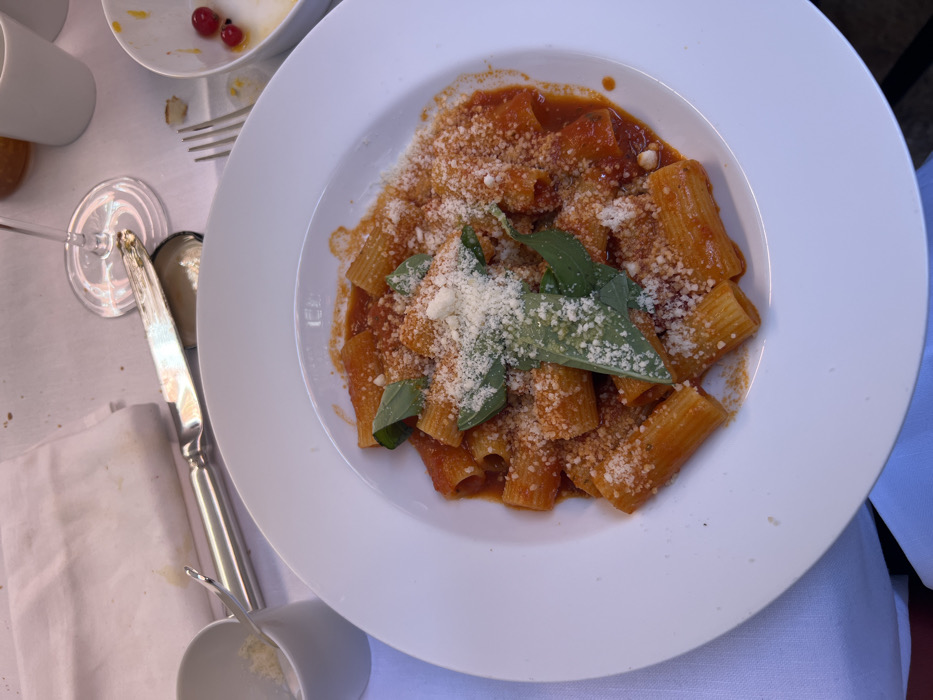 The image displays a close-up, elevated view of a dining table setting, depicting a meal within the city of Paris, France. The specific establishment or precise neighborhood is not discernible from the visual information.
The central subject is a white, wide-rimmed plate holding a portion of rigatoni pasta in a red tomato sauce. The pasta is generously topped with grated white cheese and garnished with multiple fresh green basil leaves.
To the upper left of the main plate, positioned on a white tablecloth, a small white ceramic cup contains additional grated white cheese, along with a small metal spoon. A folded white cloth napkin, exhibiting minor stains, lies adjacent to the cup. A silver-colored knife with a detailed handle is placed horizontally across the tablecloth.
To the upper right of the pasta plate, a partially consumed white bowl contains several red berries, identified as currants, alongside a small piece of bread or pastry crumb. A metal fork is visible near this bowl. The base and lower stem of a clear wine glass are also present.
The table is covered by a white tablecloth, showing some creases and minimal discoloration. No individuals are visible in the frame, but the arrangement of food and cutlery indicates an active dining scenario.