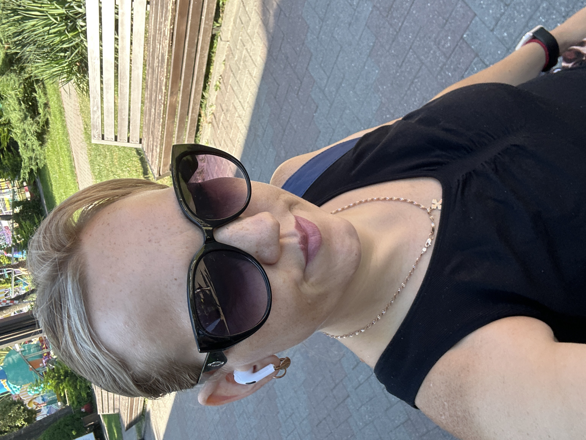A selfie taken outdoors in Gelendzhik, Russia shows a woman with short blonde hair and light skin. She is wearing black sunglasses, a black sleeveless top, and a rose gold necklace with a cross pendant. She has white earbuds in her ears and small hoop earrings. Her left hand is visible, wearing a watch with a black band and red trim. In the background, a wooden park bench is visible on a paved walkway, with trees and a playground in the distance. The lighting suggests it is daytime.