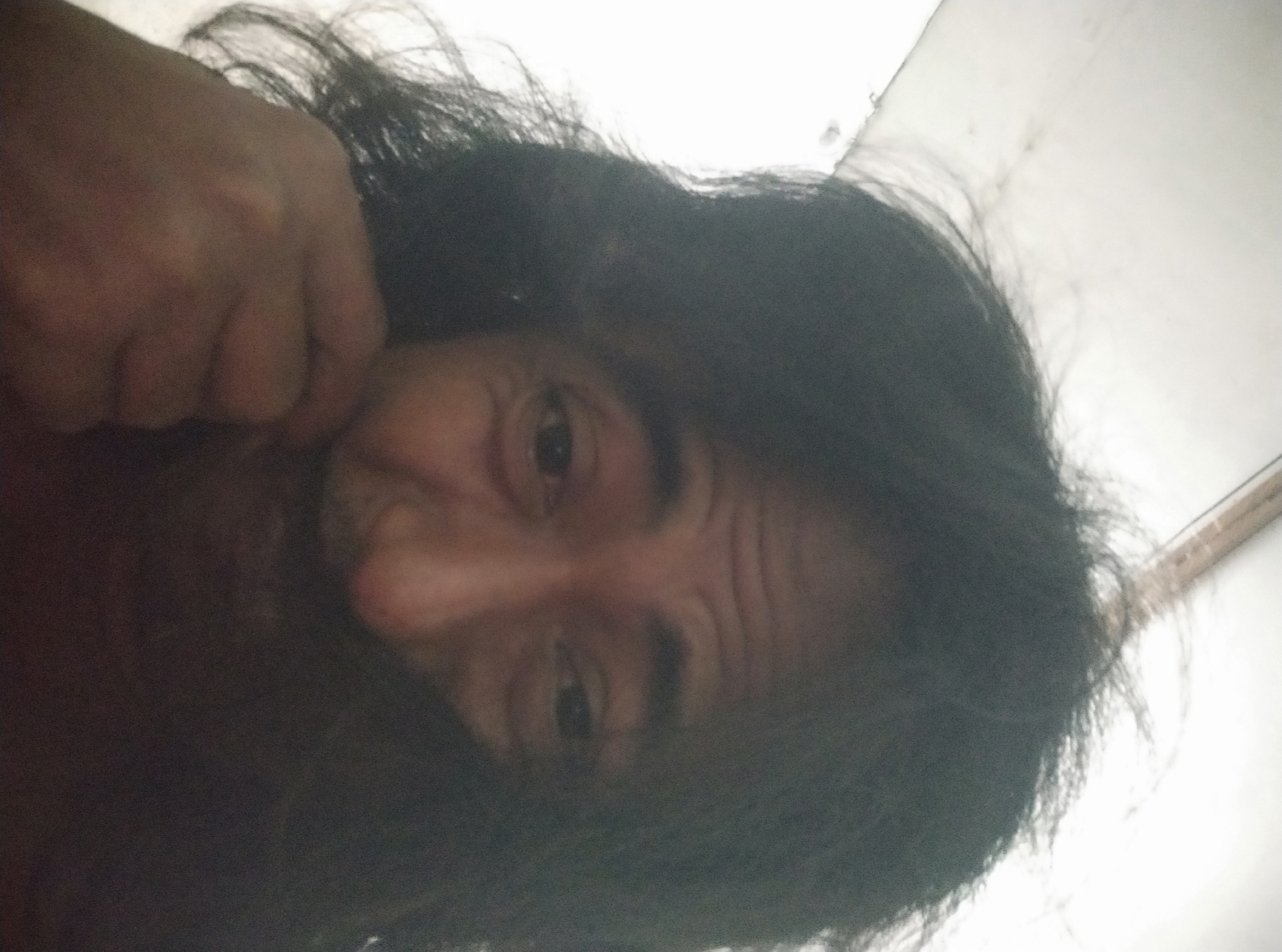 The image is a close-up, low-angle selfie of a person with long, dark, messy hair. The person's face is partially obscured by their hair and a hand, but their eyes, forehead, and part of their nose are visible. The lighting is bright, casting some shadows on the face. The background appears to be a white, textured surface, possibly a ceiling or wall. There are no identifiable landmarks or context to suggest a location in Sumurber, Indonesia.