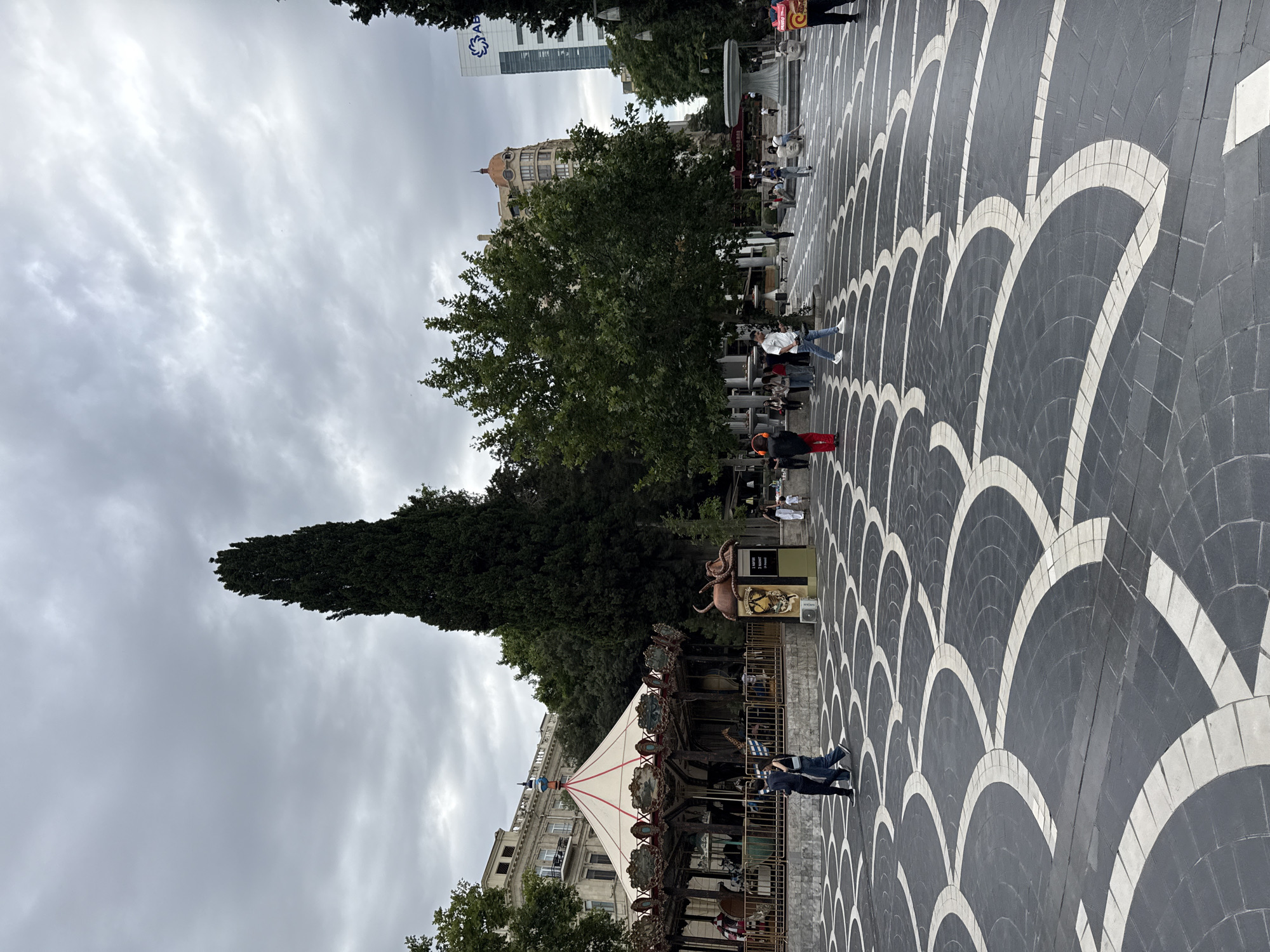 The image shows a public square with a patterned pavement. In the foreground, a carousel with a white canopy and decorative elements is visible, with several people, including children, on it. The carousel is surrounded by a fence, and there are also people standing around it.
In the background, there are trees, buildings, and a tall skyscraper with a logo. The sky is overcast and appears to be cloudy, suggesting it might be a gloomy day. The overall scene depicts a lively public space, likely a park or a recreational area, with families enjoying themselves at the carousel.
The pavement has a striking design composed of dark grey and white arched shapes, creating a dynamic visual effect. The location appears to be a city, given the presence of tall buildings and the urban infrastructure. The mood seems relaxed and joyful, as indicated by the activity of people, especially children, on the carousel.
There is some text visible on a sign near the carousel, but it is too small and blurry to read. A logo is visible on the skyscraper in the background, but its details are also unclear. The time of day is difficult to ascertain due to the overcast sky, but it appears to be daytime.