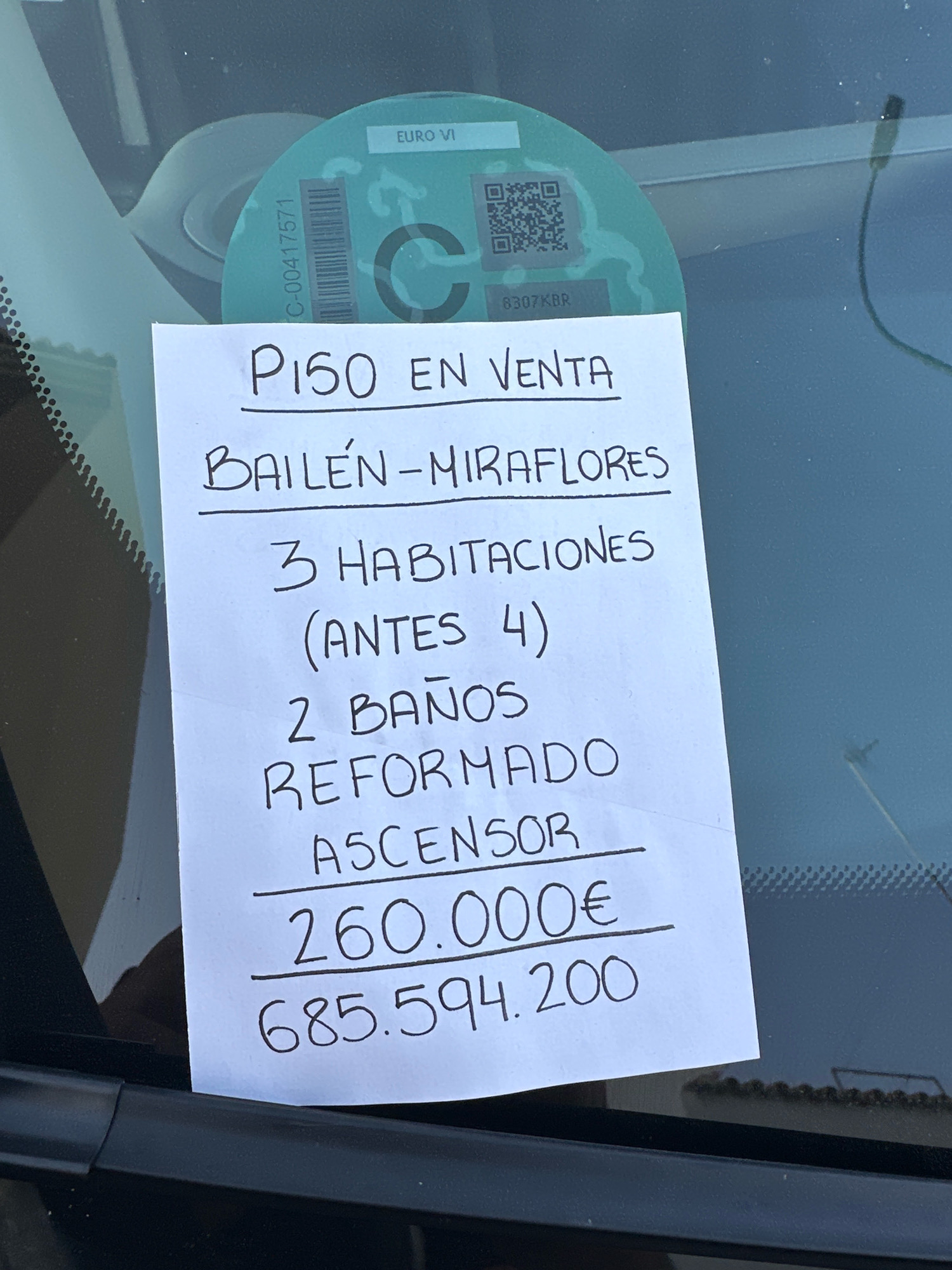 A handwritten sign taped to the inside of a car windshield advertises an apartment for sale. The sign reads "PISO EN VENTA" (Apartment for Sale) above the location "BAILÉN-MIRAFLORES". Below, it lists features: "3 HABITACIONES (ANTES 4)" (3 bedrooms (formerly 4)), "2 BAÑOS" (2 bathrooms), "REFORMADO" (Renovated), and "ASCENSOR" (Elevator). The price is listed as "260.000€", with a second figure "685.594.200" below it, which appears to be a different currency or a typo. A green environmental sticker with "EURO VI" is visible on the windshield behind the sign, partially obscuring a QR code and the number "8307KBR". The car is parked outdoors, with glimpses of a building's roofline in the reflection on the windshield, suggesting an urban environment like Málaga, Spain. The image is taken from a slightly low angle, looking up at the windshield.