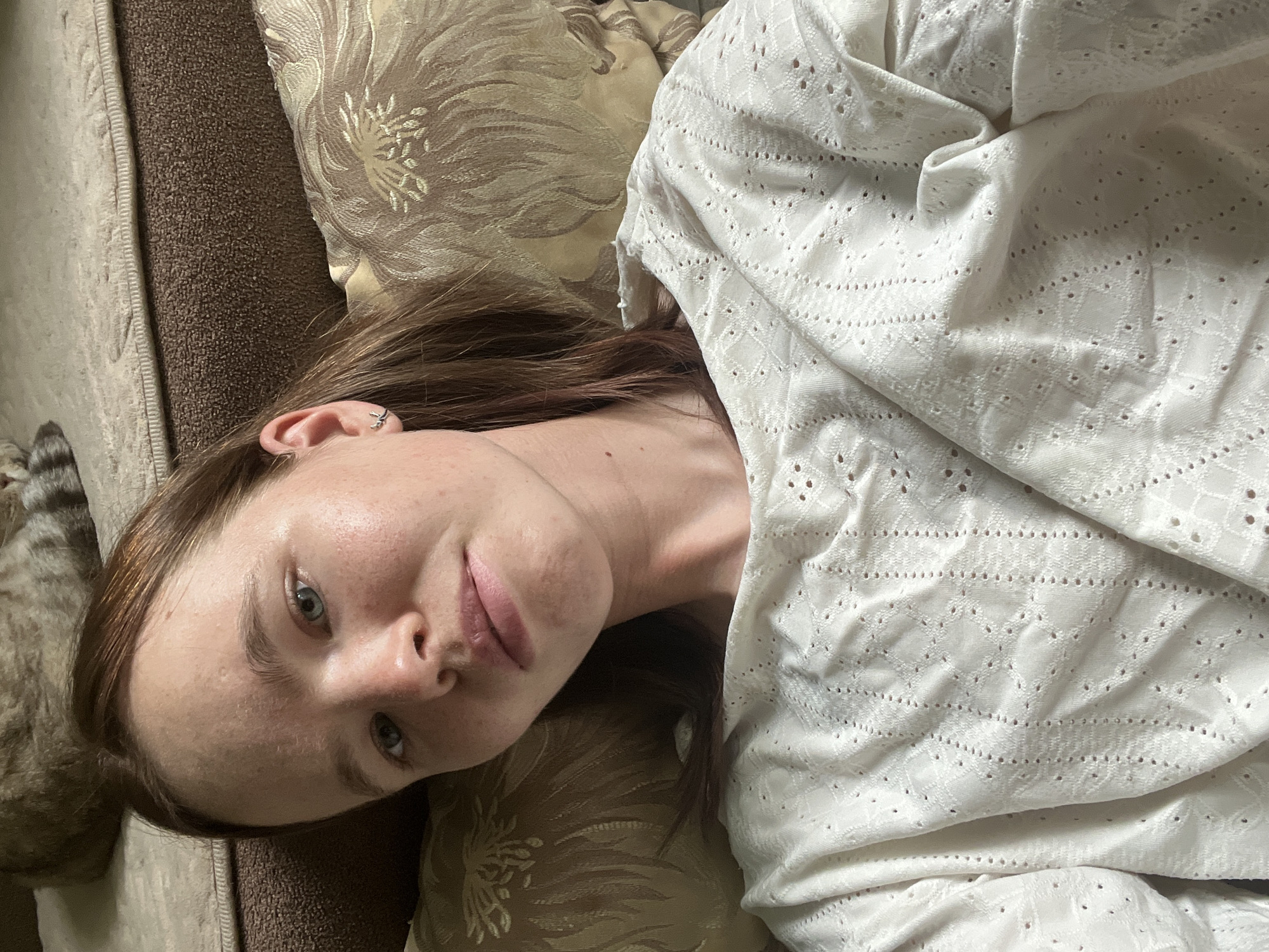 A woman is lying on a brown and beige patterned couch or bed, partially covered by a white eyelet-patterned fabric. Her head rests on a pillow. She is fair-skinned with brown hair and green eyes. She has a subtle smile. A gray cat is partially visible in the lower left corner of the frame. The location is not discernible from the provided image; the city of Chaikovsky, Russia, cannot be confirmed.