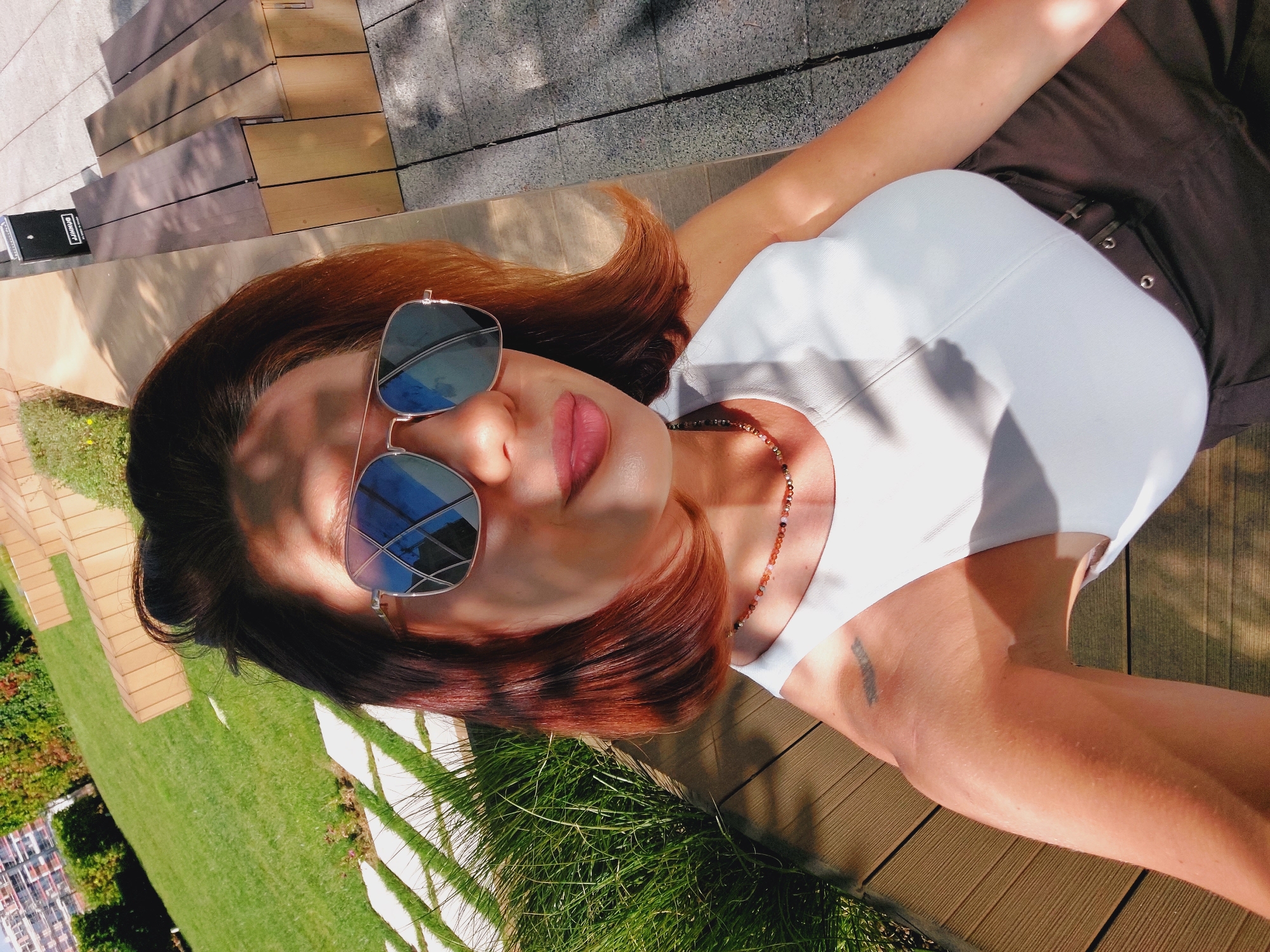 A woman with auburn hair and sunglasses is taking a selfie outdoors. She is wearing a white tank top and brown pants. The background shows a grassy area with some trees and a building. There are also wooden benches and a paved walkway with stone tiles. The scene is bathed in sunlight, creating shadows on the ground and the woman's face. The location appears to be a park or a garden within a city, possibly Sofia, Bulgaria, given the presence of modern architecture and manicured landscaping.