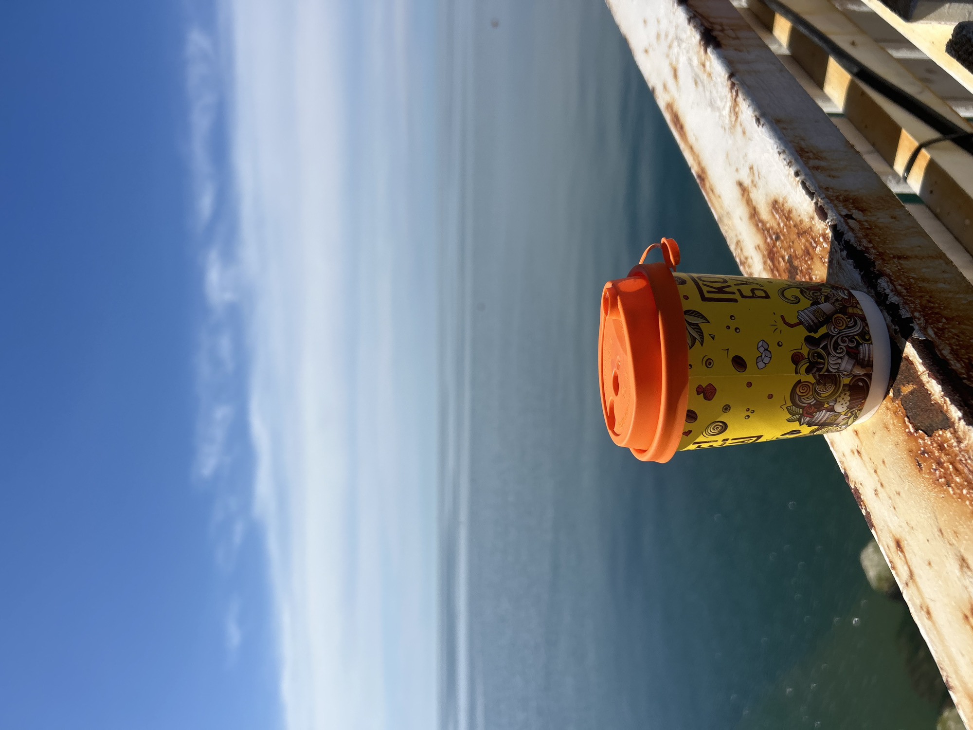 A photograph, captured outdoors in daylight, shows a yellow coffee cup with an orange lid and handle resting on a rusted metal railing against a backdrop of water and sky. The cup is partially obscured by the railing, revealing its colorful design with coffee-related illustrations. The rusted metal railing is white with brown rust and is a lattice pattern. Behind the railing, the scene transitions from a body of water to a blue sky with light clouds. The overall perspective is tilted, positioning the water and sky horizontally in the background. The location is likely Sochi, Russia.