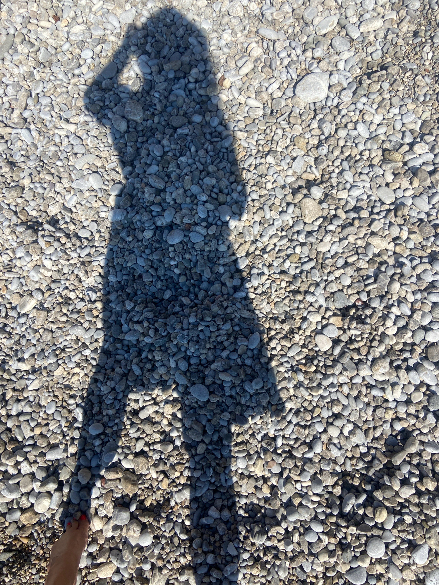 An overhead shot captures the shadow of a person standing on a beach of small, smooth, grey and white pebbles. The shadow is cast by the sun and occupies the left side of the frame, obscuring a portion of the pebbles. The person's left foot, with red nail polish, is visible at the bottom left of the image, just outside the shadow. The environment is a beach in Prijevor, Montenegro, characterized by the abundant pebbles underfoot.