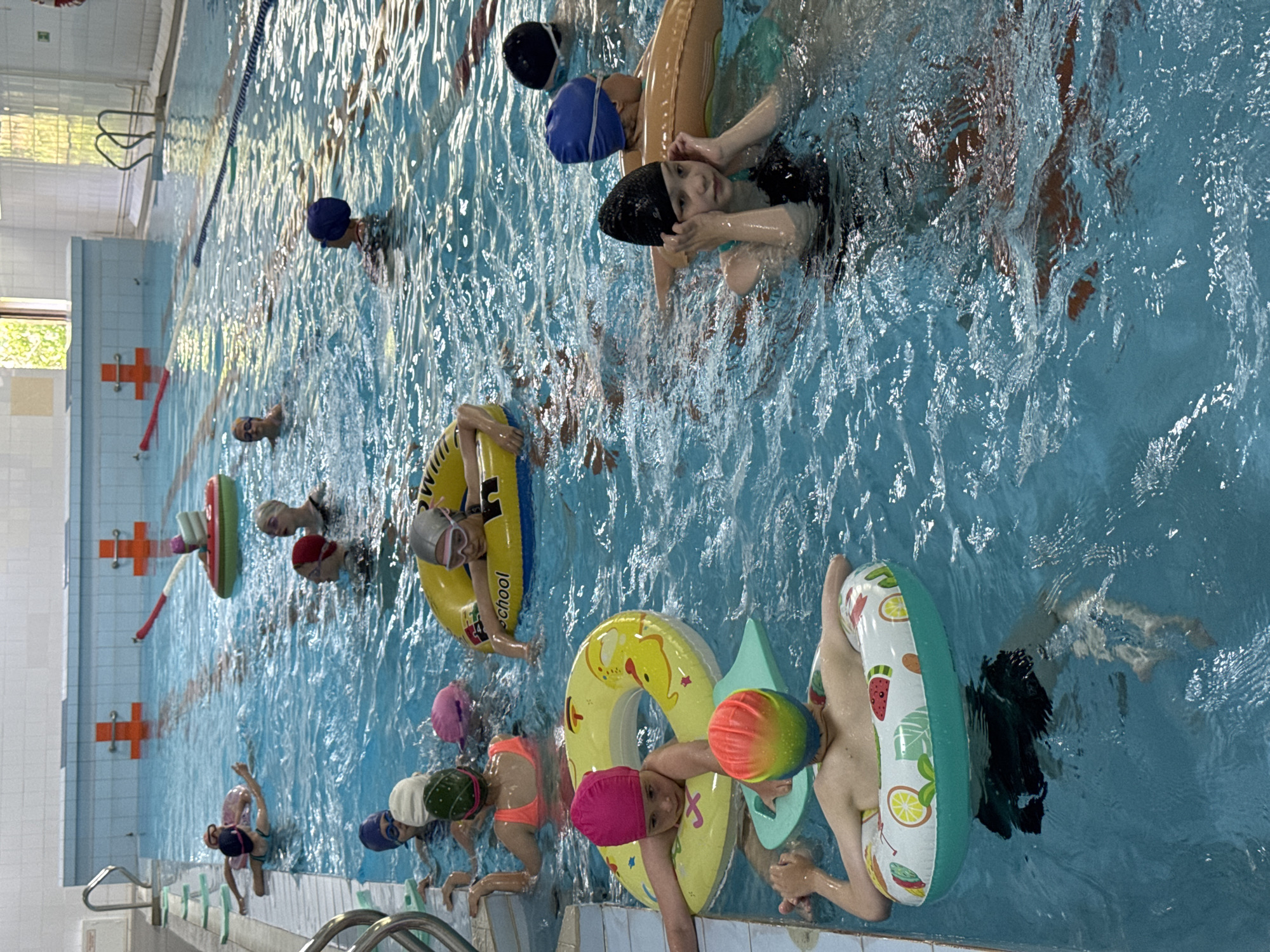 The image depicts a busy indoor swimming pool scene with numerous children participating in what appears to be a swimming lesson or recreational activity. The pool is filled with clear blue water, and lane ropes are visible, dividing sections of the pool. Several children are wearing swim caps and goggles. They are positioned in different areas of the pool, some are floating with inflatable rings, others are actively swimming or standing. Inflatable rings of various colors and designs are present, including a yellow one with the word "SCHOOL" and another with a fruit pattern. Some children are holding onto pool noodles or other flotation devices. The pool area is well-lit, with tiled walls and a high ceiling. The presence of a lifeguard chair or observation point is not evident, but the overall atmosphere suggests an organized group activity for children. The location is an indoor swimming pool. It is not possible to determine the city of Birobidzhan, Russia, or any specific geographical location from the visual information provided in the image.