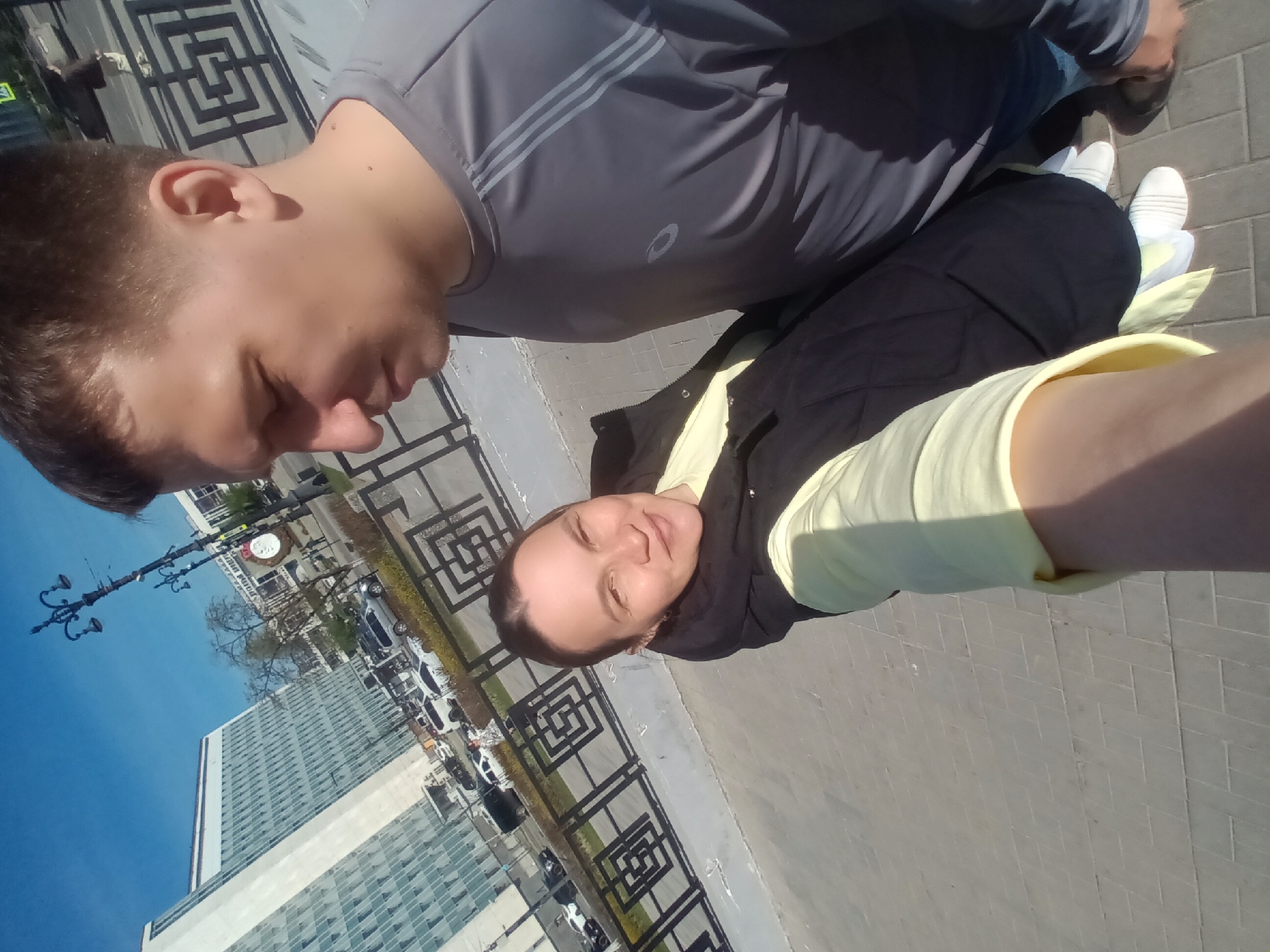 The image shows a selfie taken by a woman in Perm, Russia. She is wearing a light yellow long-sleeved shirt and a black vest. A man is also visible in the photo, wearing a gray t-shirt with stripes on the sleeves and brown shoes. Both are standing on a gray brick surface, likely a sidewalk or walkway. A black metal fence with a repeating geometric pattern is visible in the background. In the far background is a large glass building and a street with cars. A street lamp with multiple ornate lights is also present. The sky is clear and blue.