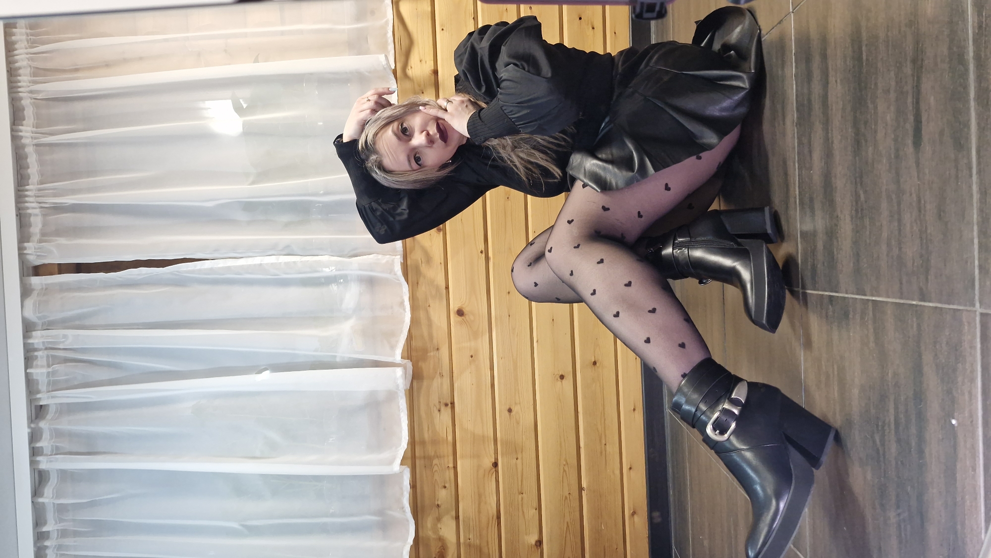 An indoor, full-body shot depicts a young woman lying on her side and posing directly at the camera. The image is oriented vertically. She appears to be in her 20s or early 30s, with light blonde hair, and a direct, slightly smiling expression.
She is dressed in a fashionable all-black outfit, featuring a black top with puffy sleeves and fitted cuffs, a black leather or faux-leather skirt, and sheer black tights adorned with small heart patterns. Her footwear consists of black ankle boots with chunky heels, a platform sole, and prominent silver buckle detailing on the straps. Her hands are positioned near her face, and her legs are crossed at the ankles.
The background on the right (when upright) consists of a warm-toned wooden wall or paneling with vertical planks. On the left, there is a white, translucent curtain or blind structure, possibly covering a window. She is lying on a dark brown tiled floor. The setting suggests an interior space, possibly a home, in Otchor, Russia. There is no visible text in the image.