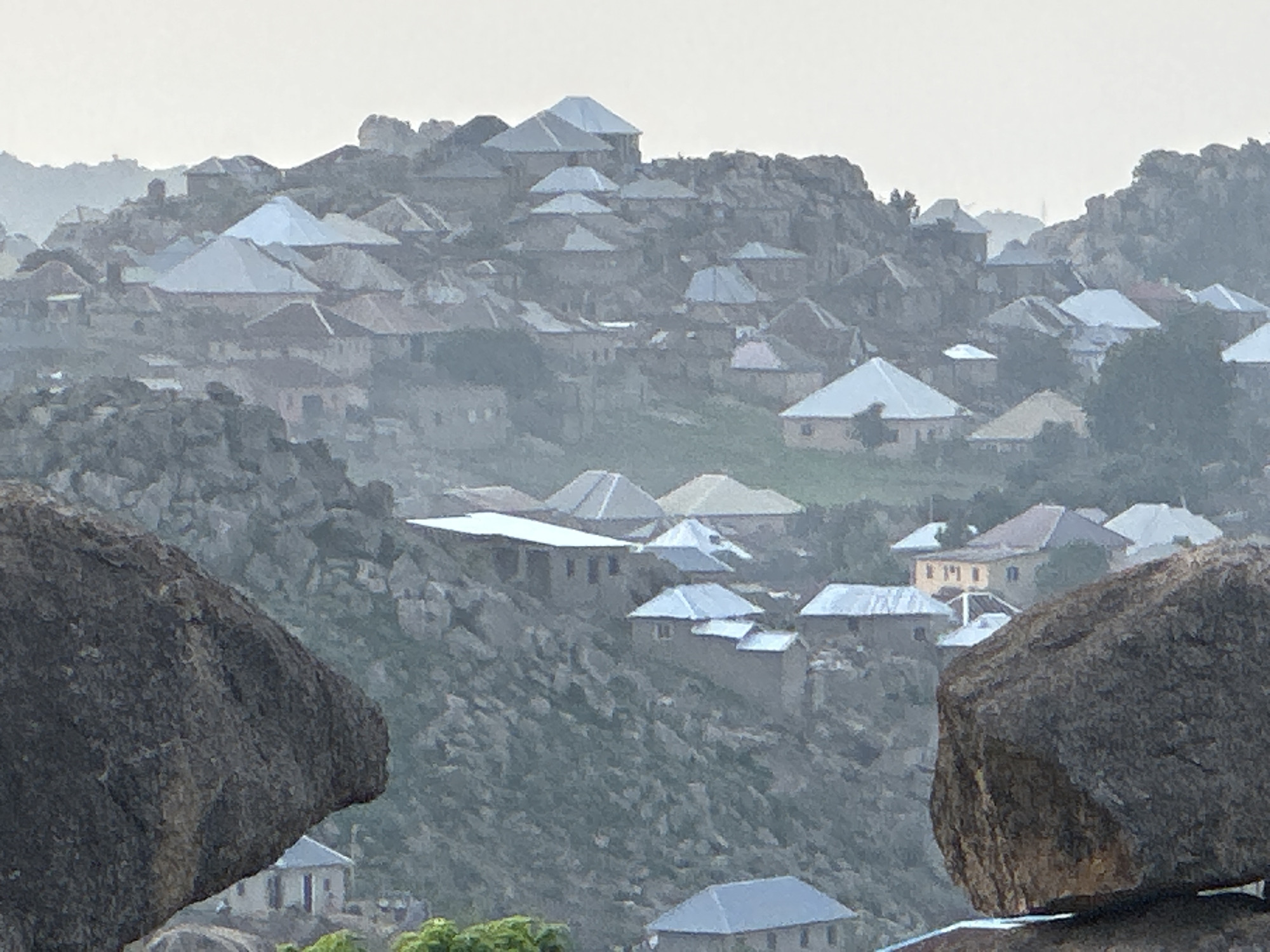 This image captures a dense village or town nestled within a prominent, rugged, and rocky landscape, characteristic of Jos, Nigeria. Numerous houses, primarily featuring light-colored, often silvery or light gray, pitched roofs, are dispersed across the undulating terrain. These structures appear to be built directly into and among massive, weathered rock formations, interspersed with sparse green vegetation. The architecture suggests an adaptation to the challenging environment, with buildings seemingly constructed from local stone or concrete materials, blending with the natural surroundings.
In the immediate foreground, two large, dark, rounded rock outcrops frame the scene, emphasizing the raw geological features that define the settlement. The houses are scattered across various elevations, some perched on hilltops, others tucked into valleys, showcasing a close integration with the natural contours of the land.
The lighting is soft and diffused, suggesting either an overcast day or the atmospheric conditions of early morning or late afternoon. A visible haze or mist creates a muted color palette dominated by grays, greens, and the pale tones of the roofs, lending a serene and somewhat distant quality to the scene. No human activity or visible text is present, allowing the focus to remain on the unique architectural and geological interplay.