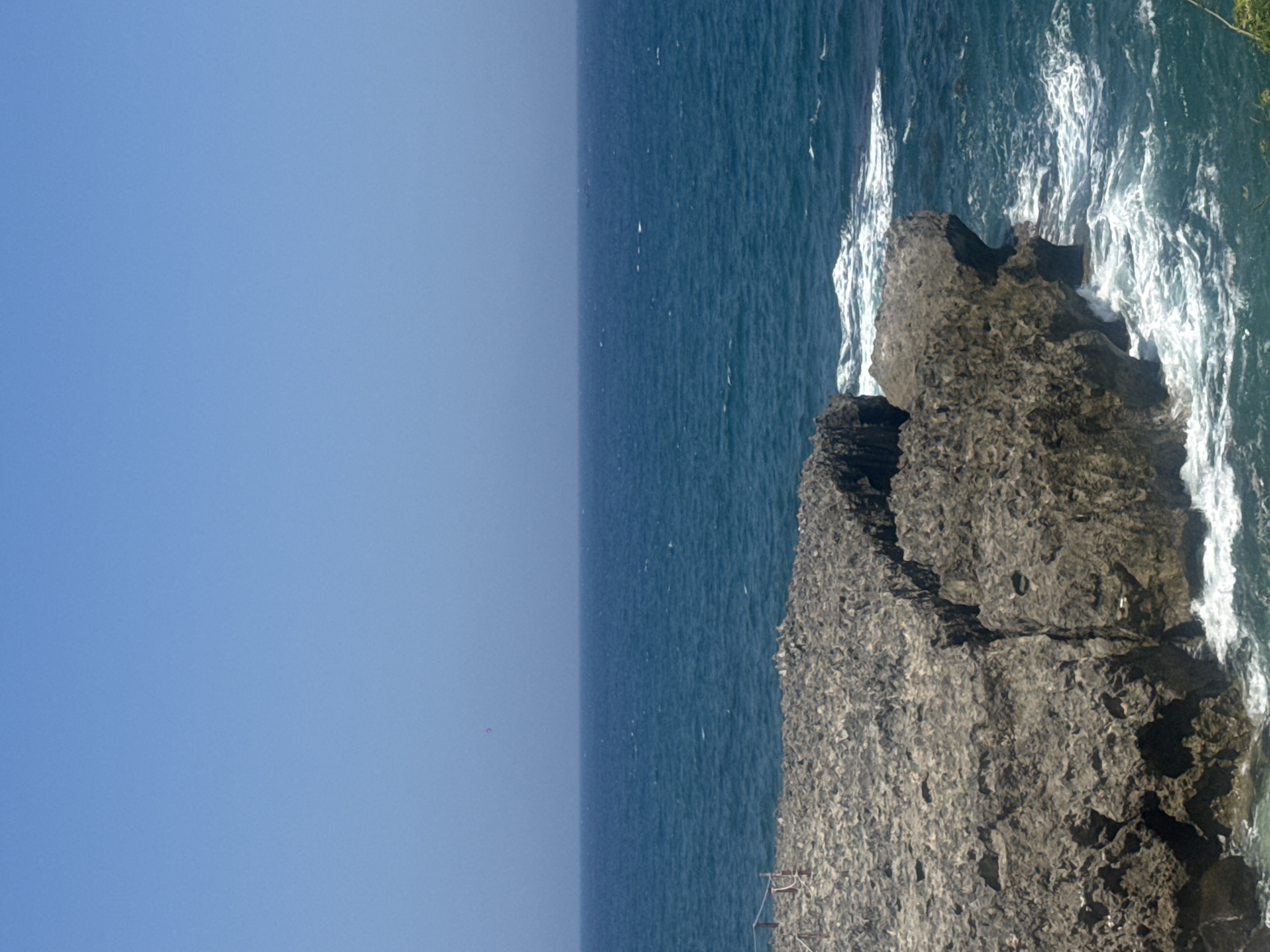 The image shows a rocky coastline with the ocean and a clear blue sky. In the foreground, there are large, rough, gray rocks that are partially submerged in the water. Waves are crashing against the rocks, creating white foam. In the background, the deep blue ocean extends to the horizon, meeting a pale blue sky. The scene appears to be a natural coastal landscape. There is no indication of human activity beyond what might be inferred from the presence of a rope barrier on the rocks. The scene is consistent with a coastal area like Vinh Hai, Vietnam, known for its rocky shores and ocean views.