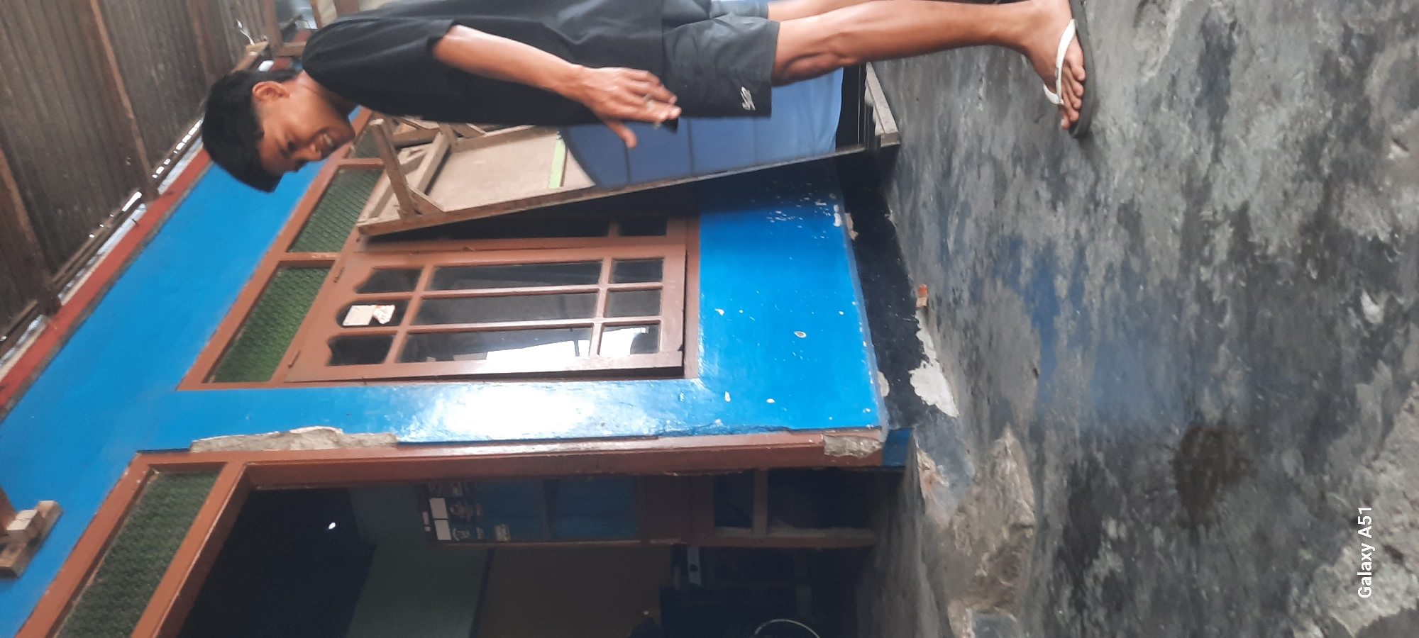 In Selopuro, Indonesia, a young male stands facing right, wearing a black t-shirt and black shorts. He is barefoot, wearing flip-flops. He is standing on a grey, weathered concrete surface. Behind him, a blue wall with wooden window frames is visible. A portion of a wooden door is also visible to the left. The scene appears to be outdoors, possibly in a residential or commercial area.