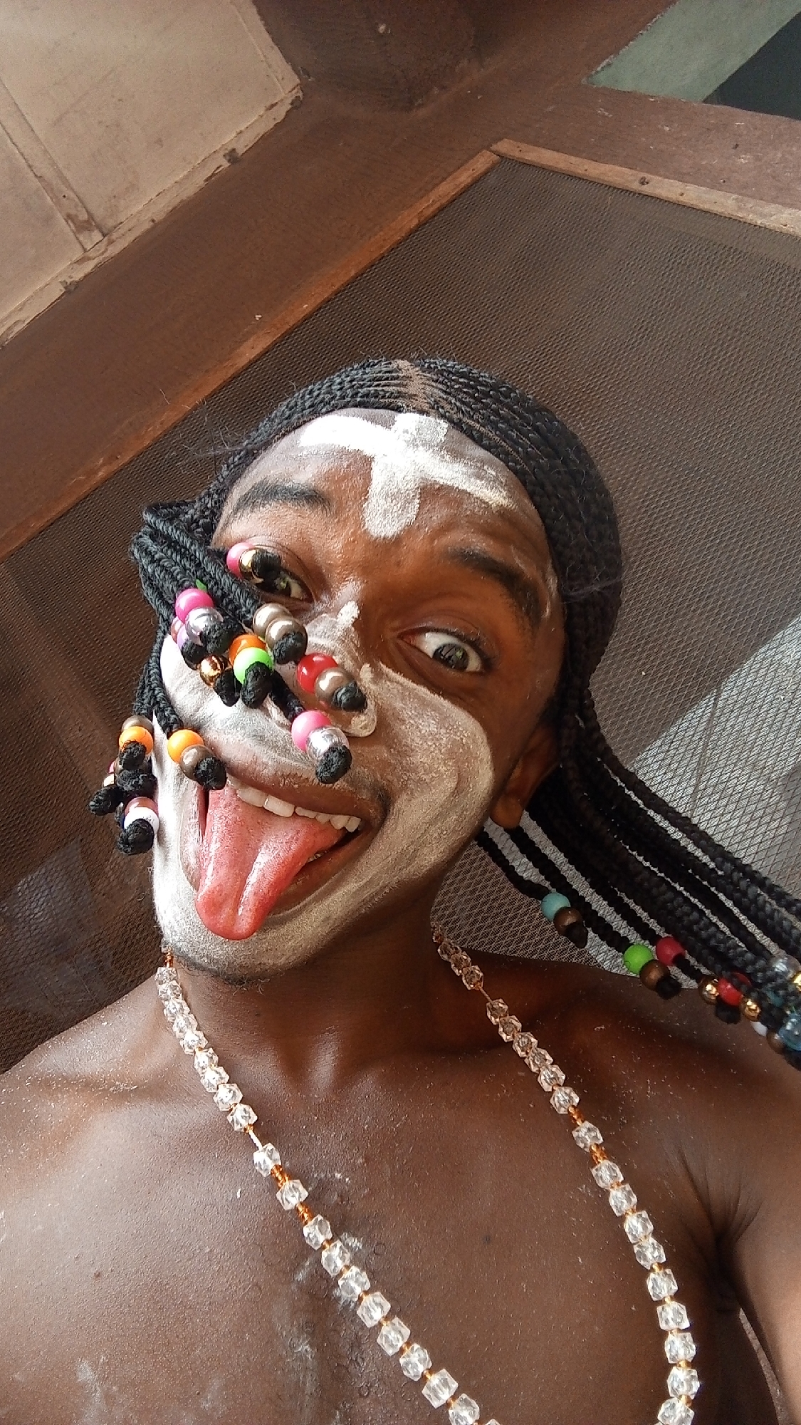 A close-up shot of a dark-skinned person with braided hair and a white substance painted on their face. The person has a white cross drawn on their forehead. They are wearing a necklace of clear, faceted beads with gold connectors. Some of the braids have colorful beads attached. The person is sticking out their tongue. The background appears to be a textured surface, possibly a screen. While it is not possible to verify a specific location without additional information, the prompt states this image is from Onitsha, Nigeria.