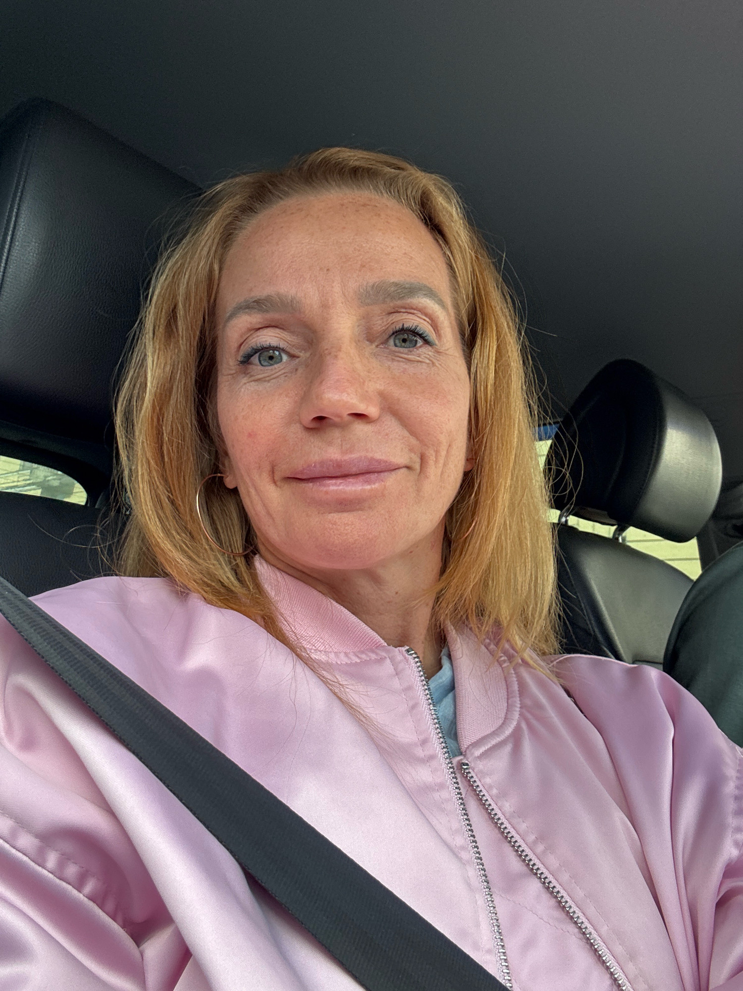 The image is a selfie taken inside a car, featuring a woman with blonde hair and light blue eyes. She is wearing a light blue t-shirt and a pink bomber jacket. A black seatbelt is visible across her chest. The woman has freckles on her face and is wearing hoop earrings. The background shows the interior of the car, including black leather headrests and a hint of the outdoors through the car window. There is no identifiable information within the image to suggest a specific location in Saint Petersburg, Russia.