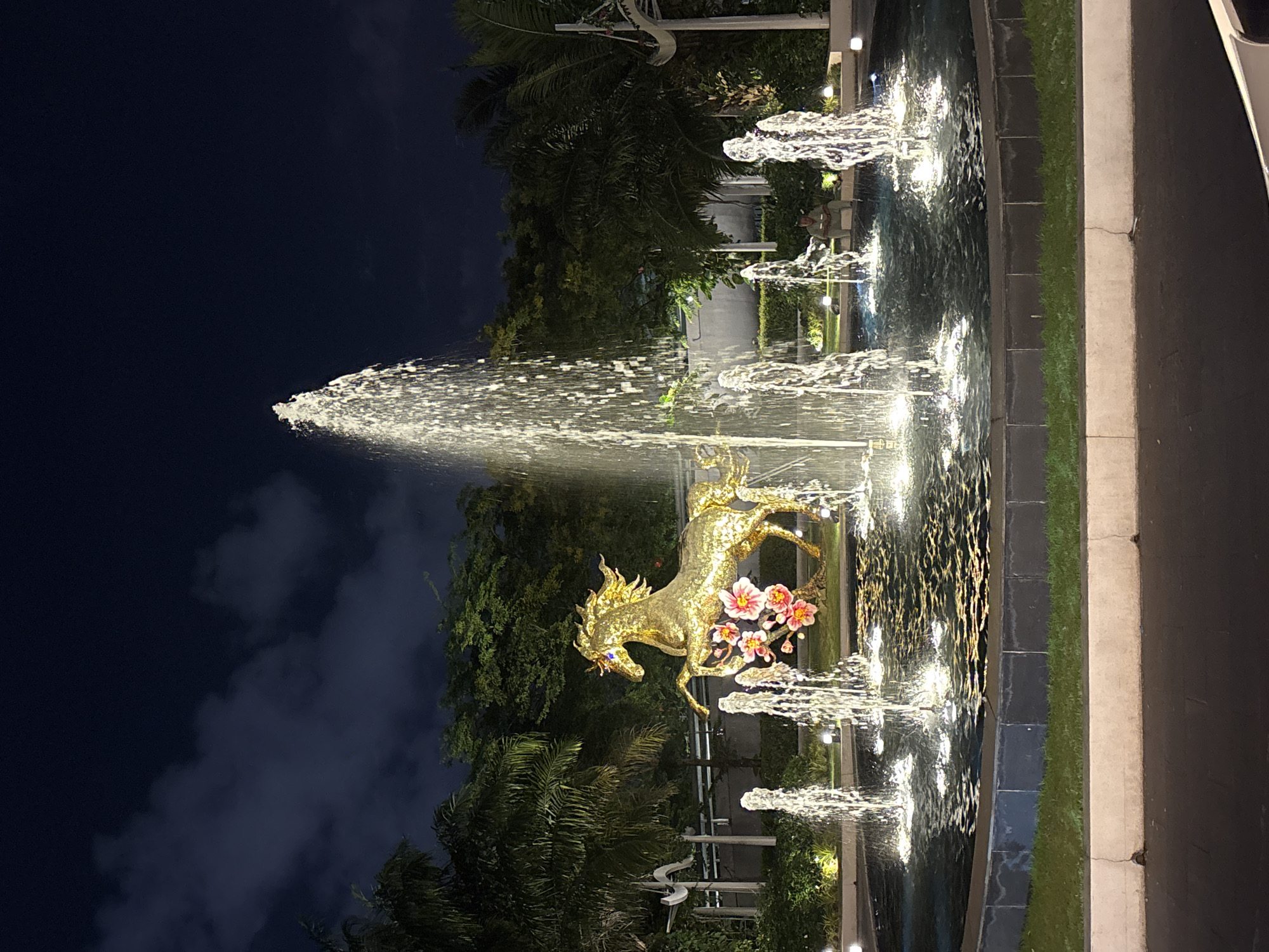 The image shows a nighttime view of a fountain featuring a golden horse statue. The horse is adorned with pink flowers. Water jets illuminate the scene. Trees and plants frame the fountain. The sky is dark with some clouds. The fountain is situated near a road or sidewalk. Based on the distinctive golden horse statue, it is likely located on đường 2 Tháng 9 in Đà Nẵng, Vietnam.