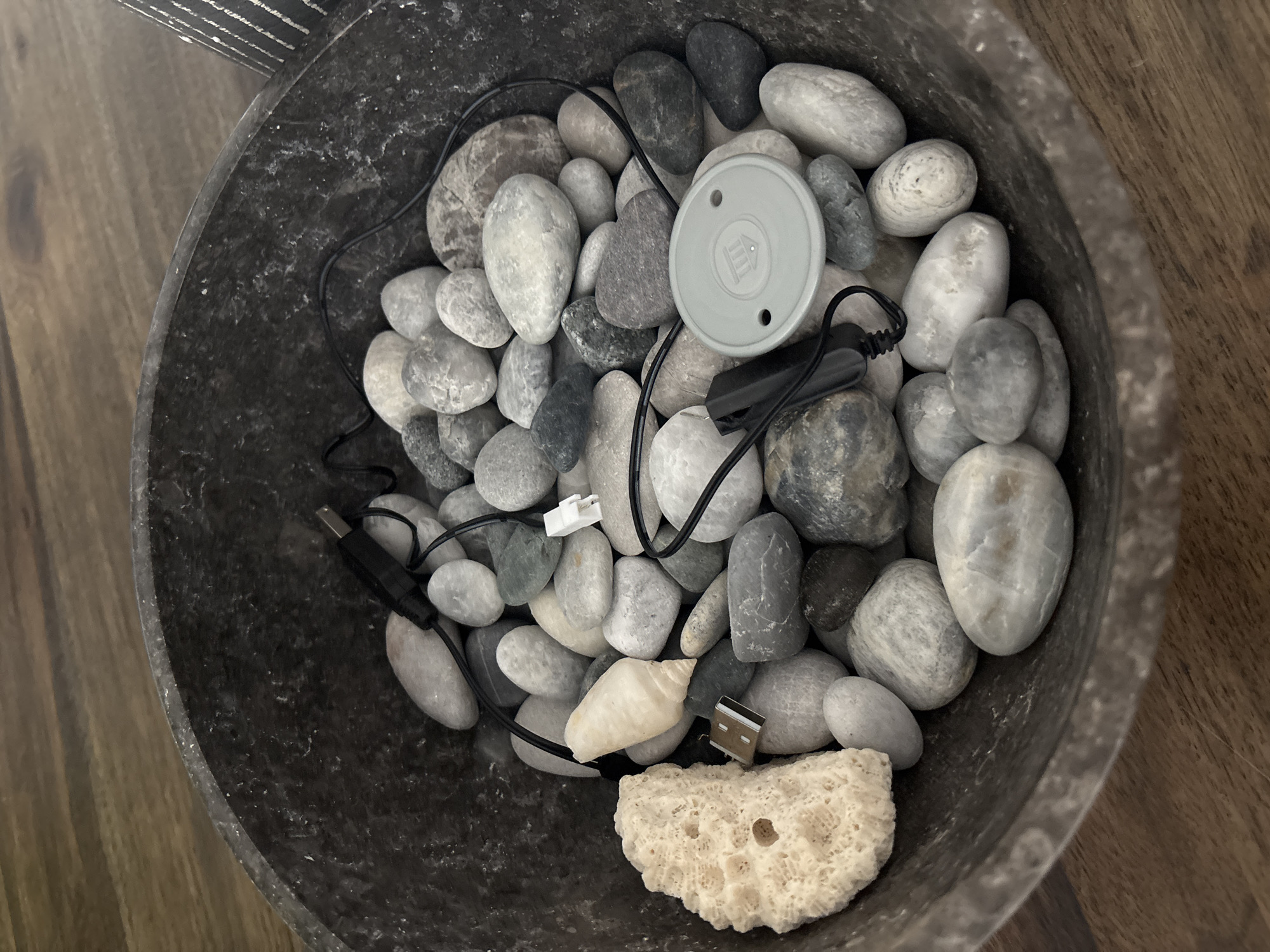 A dark, rough-textured bowl filled with smooth, grey, and white river stones is situated on a wooden surface. Among the stones are several electronic cables with USB connectors, a small white connector, and a light grey circular device with two holes and an embossed symbol. A piece of porous, light-colored coral and a small, pale seashell are also visible. The lighting is overhead, casting subtle shadows and highlighting the textures of the stones and bowl. The environment appears to be indoors, with the wooden surface suggesting a table or countertop. There is no indication of a specific location in Dubai, United Arab Emirates.