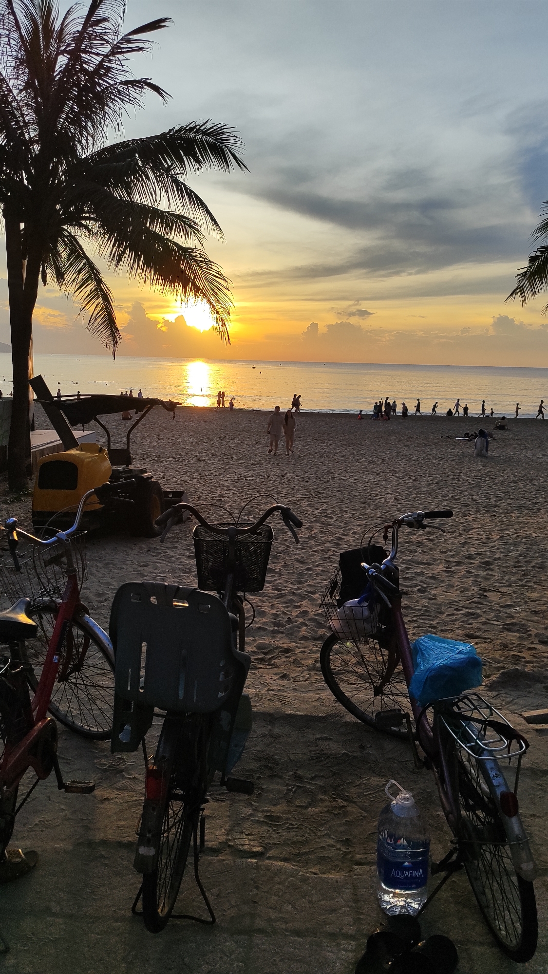 The image depicts a beach scene at sunrise in Đà Nẵng, Vietnam, likely near a populated area given the presence of people and a yellow construction vehicle. In the foreground, three bicycles are parked on the sand, two of which have baskets. One bicycle on the right has a blue plastic bag tied to its rear rack, and a bottle of Aquafina water is placed on the ground next to it. A pair of black flip-flops are also visible. The sand is textured with footprints and tire tracks. In the middle ground, several people are walking and standing on the beach. The sea is calm with a reflection of the sun on its surface. The sky is a gradient of orange, yellow, and gray, with scattered clouds, indicating the sun is low on the horizon. Two palm trees frame the left side of the image. The overall atmosphere is peaceful and serene, characteristic of a sunrise on a beach.