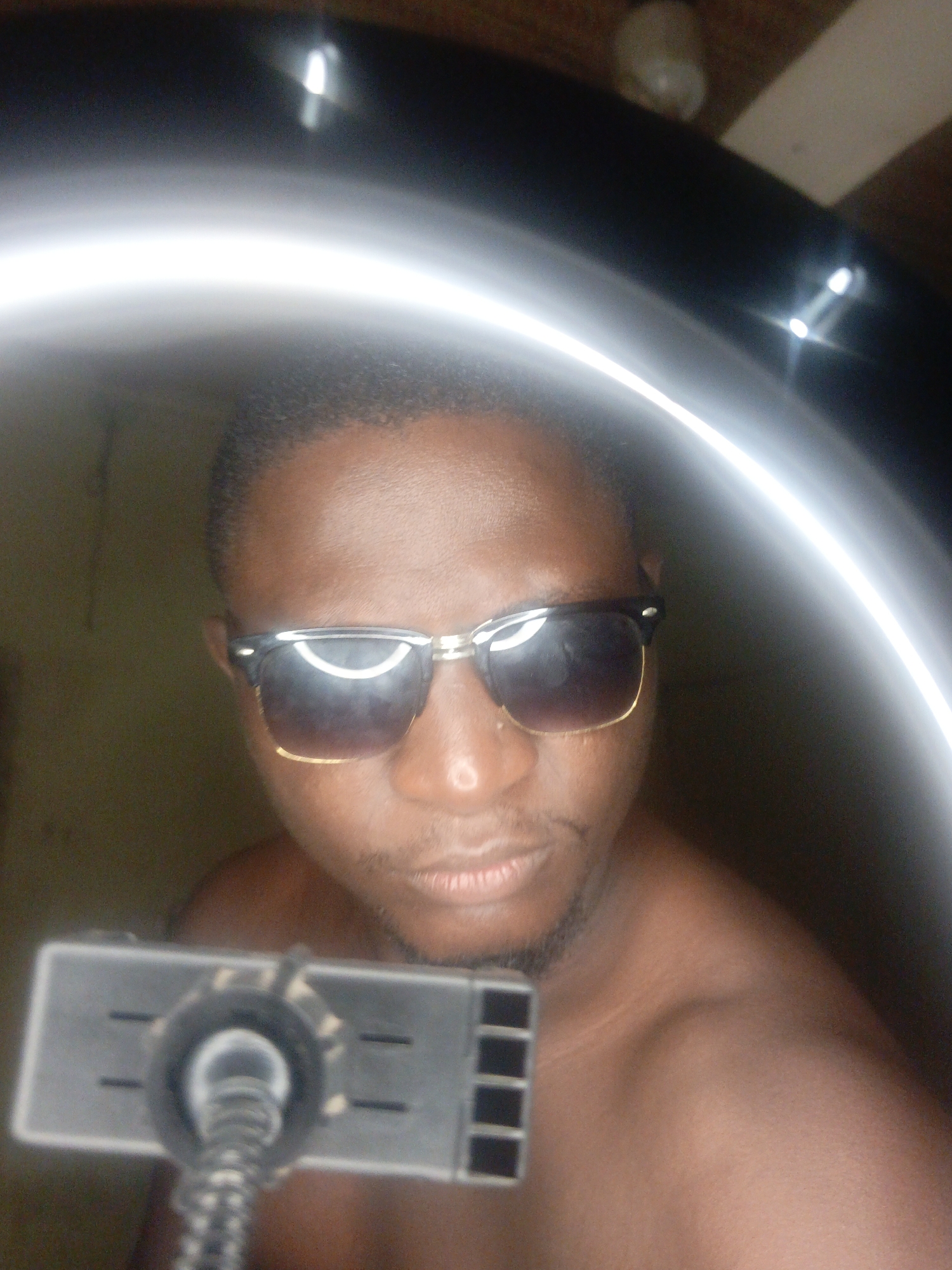 The image shows a close-up, waist-up view of a young Black person wearing sunglasses. They are looking directly at the camera, with the upper part of their body and face filling most of the frame. The person has short hair and appears to be shirtless. They are wearing dark sunglasses with a gold rim. In the foreground, a device with a coiled cable and rectangular slots is visible, positioned at chest level. The background is dark and indistinct, with a bright, circular light source that creates a halo effect around the subject's head. There are no discernible details about the location within a city.