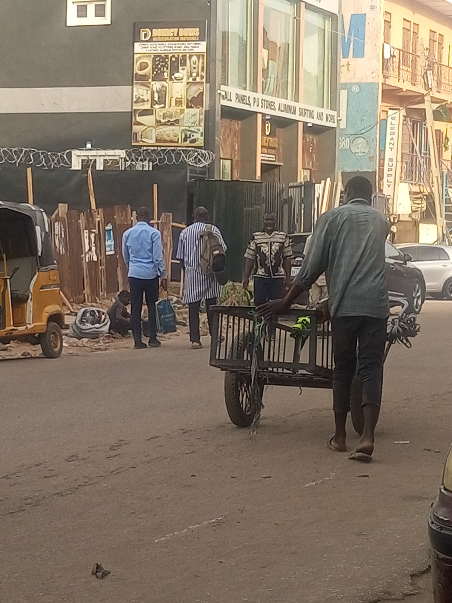 The image shows a street scene in Maiburiji, Nigeria. A man pulls a wooden cart loaded with green coconuts. Three other men walk in the same direction. A yellow auto rickshaw (tuk-tuk) is parked on the side of the street. One man is sitting on the ground next to the rickshaw. Various businesses are visible in the background, including "Domict Bums," which sells "PERICEBATION MATERIALS," and a store advertising "BALL PANELS, PU STONES ALLUMINIUM SORTING AND WORE." "CLEBANT SUPPLY" is also visible on a sign. A black car is parked further down the street. The sky appears overcast.
