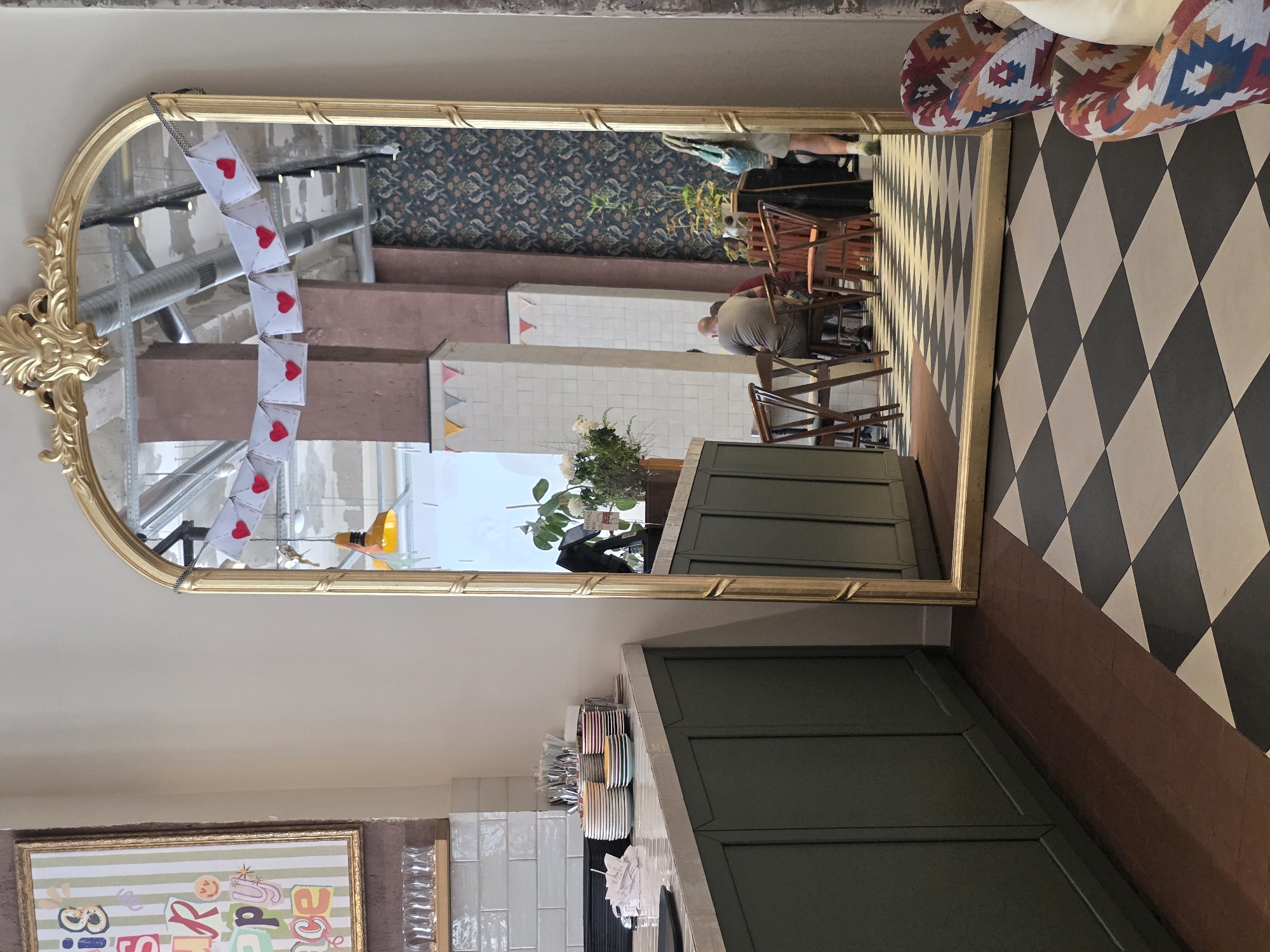 The image depicts the interior of what appears to be a cafe or restaurant. A large, ornate gold-framed mirror dominates the center of the scene, reflecting the opposite side of the room. Above the reflection of the cafe's seating area, a string of white envelopes adorned with red hearts hangs, suggesting a celebratory or romantic theme. The cafe's interior features a black and white checkered floor, dark green cabinetry along the left wall, and stacks of colorful plates on the counter. In the reflection, several tables with wooden chairs are visible, with a few patrons seated, including a person seen from behind wearing a grey t-shirt. The architecture includes exposed pipes and industrial-style lighting. A vibrant, framed artwork with colorful lettering is partially visible on the left wall, and to the right of the mirror, two upholstered chairs with a patterned fabric are positioned. The overall impression is a well-lit, contemporary space with decorative elements. No specific landmarks or indications of Khatay Raion, Azerbaijan are present in the visual information.