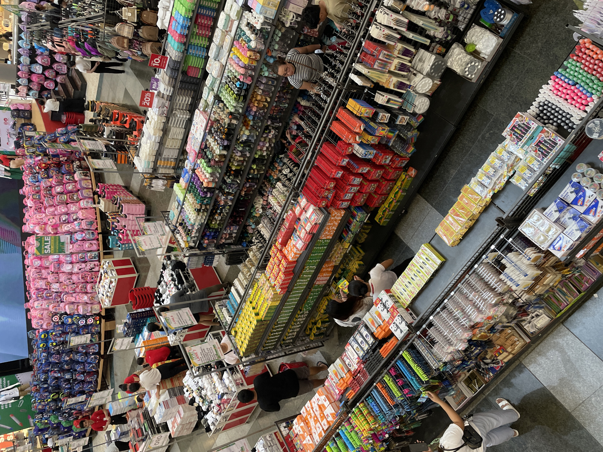 This is an overhead shot of a busy department store, likely located in Davao City, Philippines, judging by the presence of "SM Store" branding visible on some of the signage. The scene is filled with a wide assortment of merchandise, primarily school and art supplies.
In the foreground, a woman in a white top and grey pants is examining items displayed on a shelf. To her right, a conveyor belt is visible, with several rows of packaged goods, including what appear to be art supplies like markers and crayons. Further to the right, a man in a beige shirt is looking at products.
The mid-ground reveals more shelves stocked with various items. Rows of colorful stationery, pens, and other school supplies are neatly arranged. Several shoppers, distinguishable by their attire, are browsing the merchandise. One woman in a striped shirt and glasses is looking at products on a shelf.
In the background, the store opens up to a much larger section displaying an extensive collection of backpacks. These backpacks are organized by color, with large sections of blue, pink, and purple bags. Many of these bags feature cartoon characters and are displayed in large quantities, suggesting a back-to-school sale. A red display bin with more backpacks is also present.
Throughout the store, promotional signage is visible, including "SALE" banners and price reductions like "20% OFF" and "LESS 10%". Staff members, some wearing red shirts, are also seen assisting customers or organizing merchandise. The overall environment is bright and organized, characteristic of a retail setting. The flooring appears to be a polished grey material.