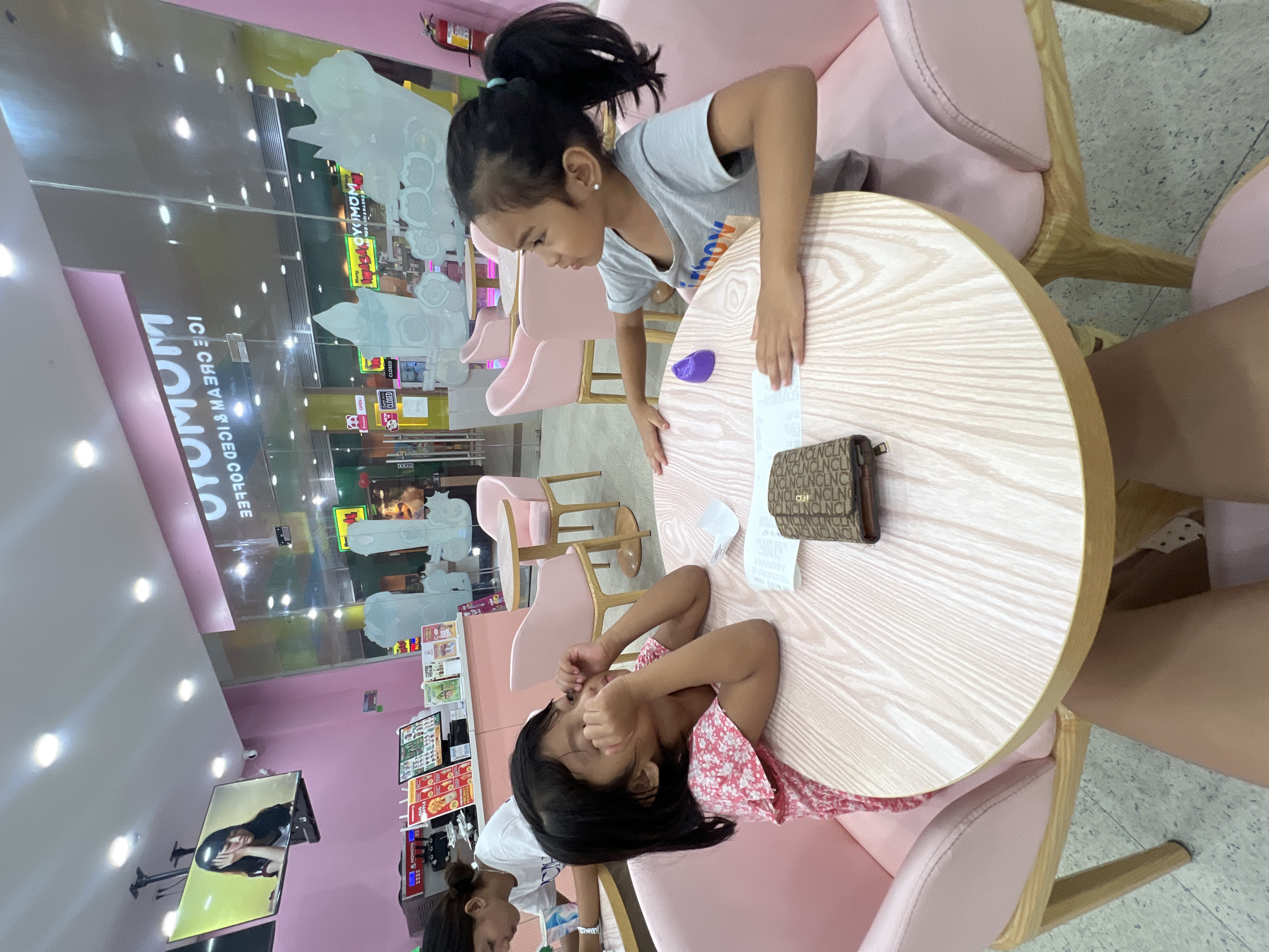 In Panabo City, Philippines, two young girls are seated at a round, light-wood-grain table inside a bright pink establishment identified by signage as "MOMOYMOM ICE CREAM & ICED COFFEE." The girl on the left, wearing a pink floral-patterned top, is leaning forward with her hands near her eyes, appearing to rub them. The girl on the right, in a grey t-shirt, is looking to her left, with her arms resting on the table. Between them, on the table, are a folded wallet with a monogrammed pattern, a white paper, and a small purple object. The chairs are upholstered in a soft pink. Behind them, through a glass partition, the interior of the establishment is visible, including more seating, menu displays, and the counter area. A large television screen mounted on the ceiling displays an image of a young woman. The overall environment suggests a casual dining or dessert shop.