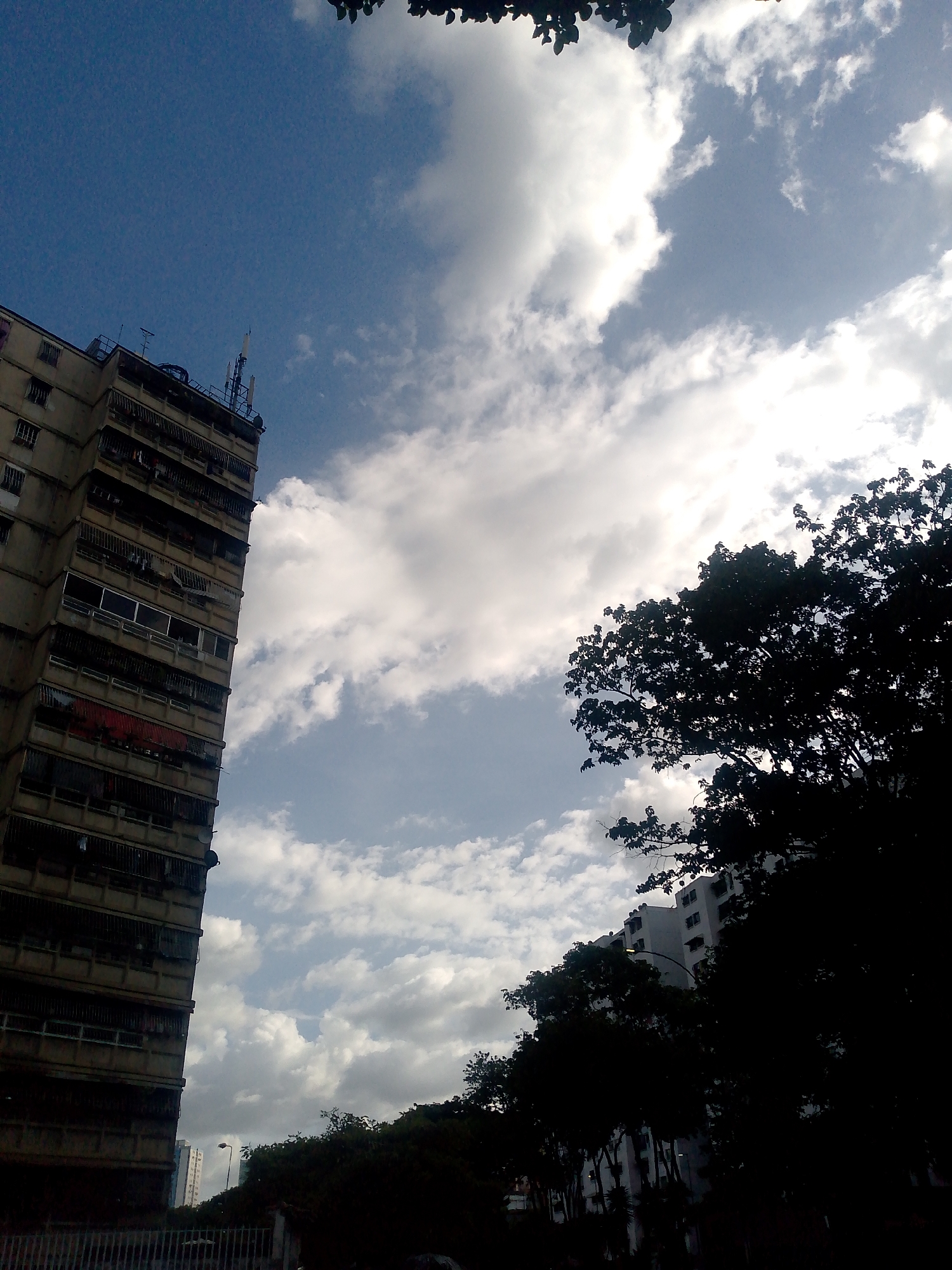 The image shows a tall, weathered apartment building on the left side, possibly in Caracas, Venezuela, as evidenced by the architecture and dense urban setting. A complex network of antennas is visible on its roof. White clouds fill the sky, with a strong sun partially obscured, creating bright and shaded areas. To the right, dark silhouettes of trees stand in the foreground, obscuring other buildings. The presence of a fence and another building further back behind the trees suggests a dense urban environment. No people are visible.