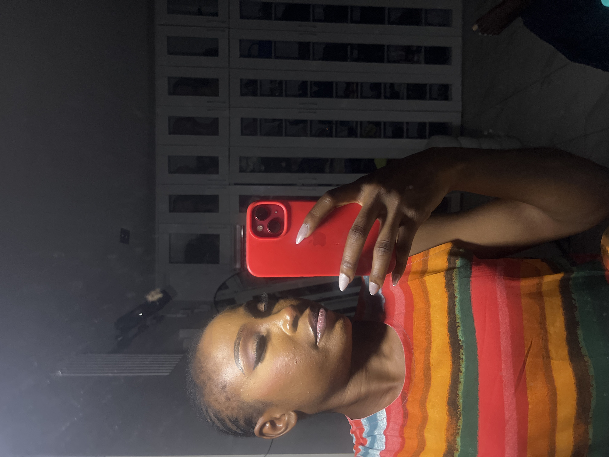 The image shows a close-up, selfie-style shot of a woman in what appears to be a well-lit room. The woman is positioned on the left side of the frame, facing slightly to the right, and is holding a bright red smartphone in her right hand, which is capturing the reflection. She has dark skin, is wearing eye makeup and lip gloss, and has her hair styled in small braids pulled back. Her attire consists of a colorful, horizontally striped t-shirt featuring shades of orange, yellow, green, and red.
The background is dominated by a large, reflective surface, likely a mirror or a glass-fronted wardrobe, which occupies the majority of the right side of the image. Behind the woman, the reflection reveals a room with what appear to be tall, white cabinets or closets with multiple glass panes, suggesting a storage area or a walk-in closet. The lighting in the room is somewhat dim, with shadows cast by the objects within the reflective surface. To the far right, in the reflection, there is a glimpse of two feet belonging to another person, indicating another individual is present in the room, though out of focus. The overall impression is that of a casual, personal moment captured within a domestic setting, possibly in a residence. There is no specific indication that this location is in Abuja, Nigeria.