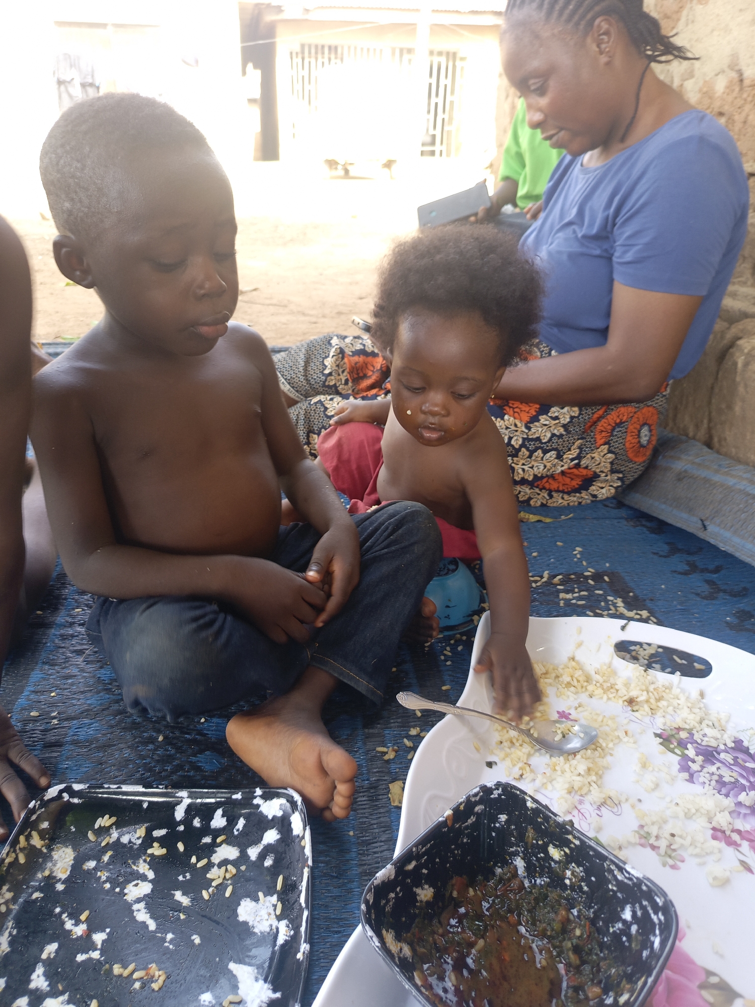 In an outdoor scene, two young children sit on a blue patterned mat, with an adult woman seated behind them to the right. The older child, a boy, is shirtless and wears denim jeans. To his right, a younger child, possibly a toddler, is also shirtless and has a small amount of food on their face. Both children appear to be focused on food placed on a white tray in front of them. A spoon rests on the tray amongst the scattered food. A dark bowl containing food is situated near the tray. Another container with remnants of food is on the left side of the mat near the older child. The adult woman is wearing a blue top and a patterned skirt. The location is Unknown, Unknown.
