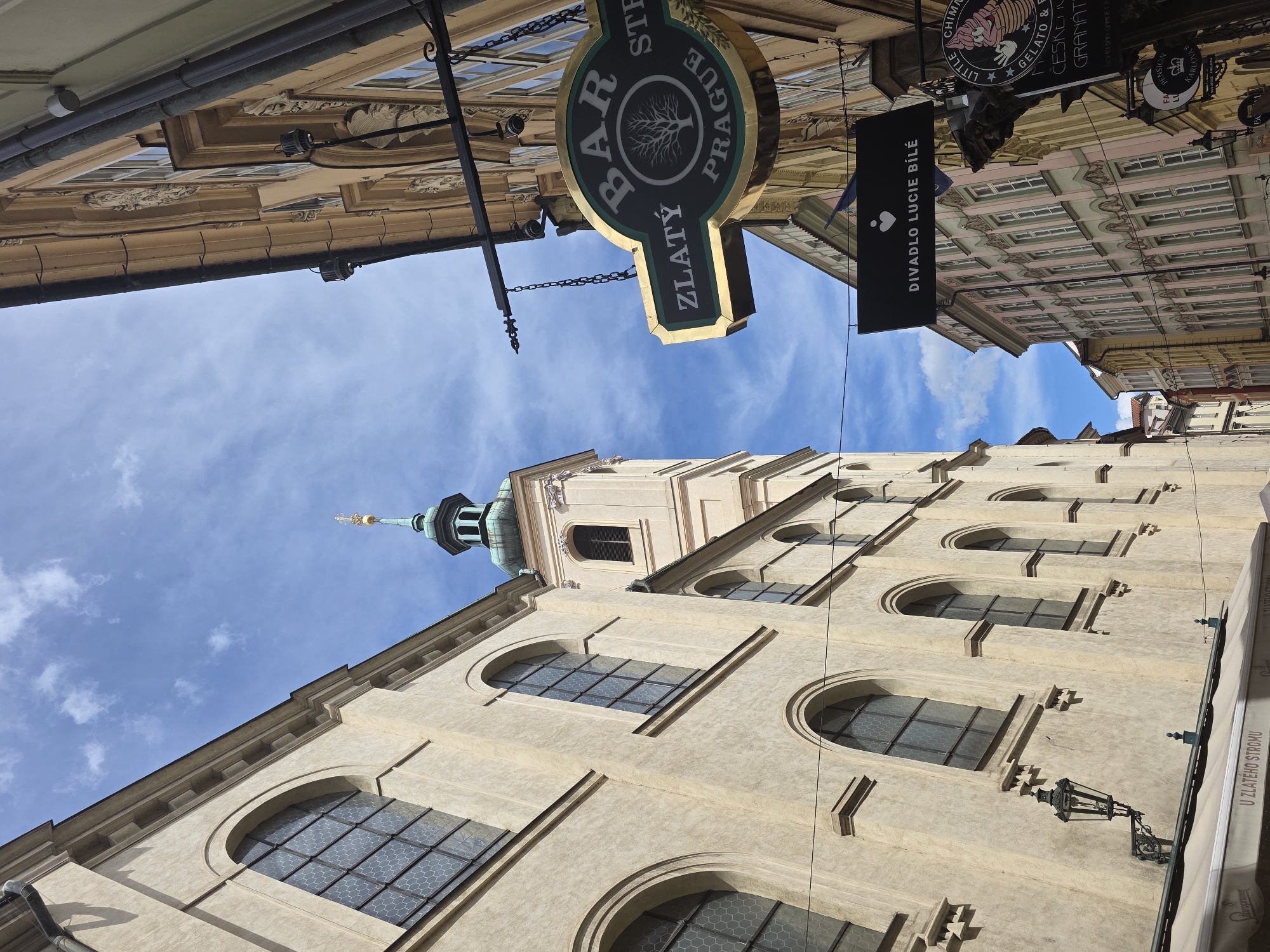 An upward-angled view captures a historic street in Prague, Czech Republic, under a bright blue sky with scattered white clouds.
On the left, a multi-story building with a light cream-colored stucco facade features a series of tall, arched windows with dark frames and a hexagonal grid pattern. Architectural details include cornices and ledges above and between the windows. A prominent light beige clock tower with a dark louvered opening is visible behind or to the right of this building, topped by an octagonal teal-colored dome with a golden finial. A black, ornate gas lamp is attached to the lower section of this building, above a cream-colored awning displaying the text "U ZLATÉHO STROMU."
On the right, a row of multi-story buildings lines a narrower street or alley. These buildings exhibit varied architectural styles with ornate facades, bay windows, and colorations including pale pink, beige, and light green. Multiple signs hang from these structures: A large, oval, dark green sign with a gold border and ornate black ironwork reads "BAR ZLATÝ" above a stylized tree emblem and "PRAGUE" below it. Directly beneath, a rectangular black sign with a white heart symbol prominently displays the text "DIVADLO LUCIE BÍLÉ." Partially visible signs further down include "LITTLE CHIMN..." (likely referring to chimney cake), "GELATO & F...", and "PENSION KARLOVA." Thin black cables traverse the upper section of the sky between the buildings. The scene appears devoid of people or overt activity.