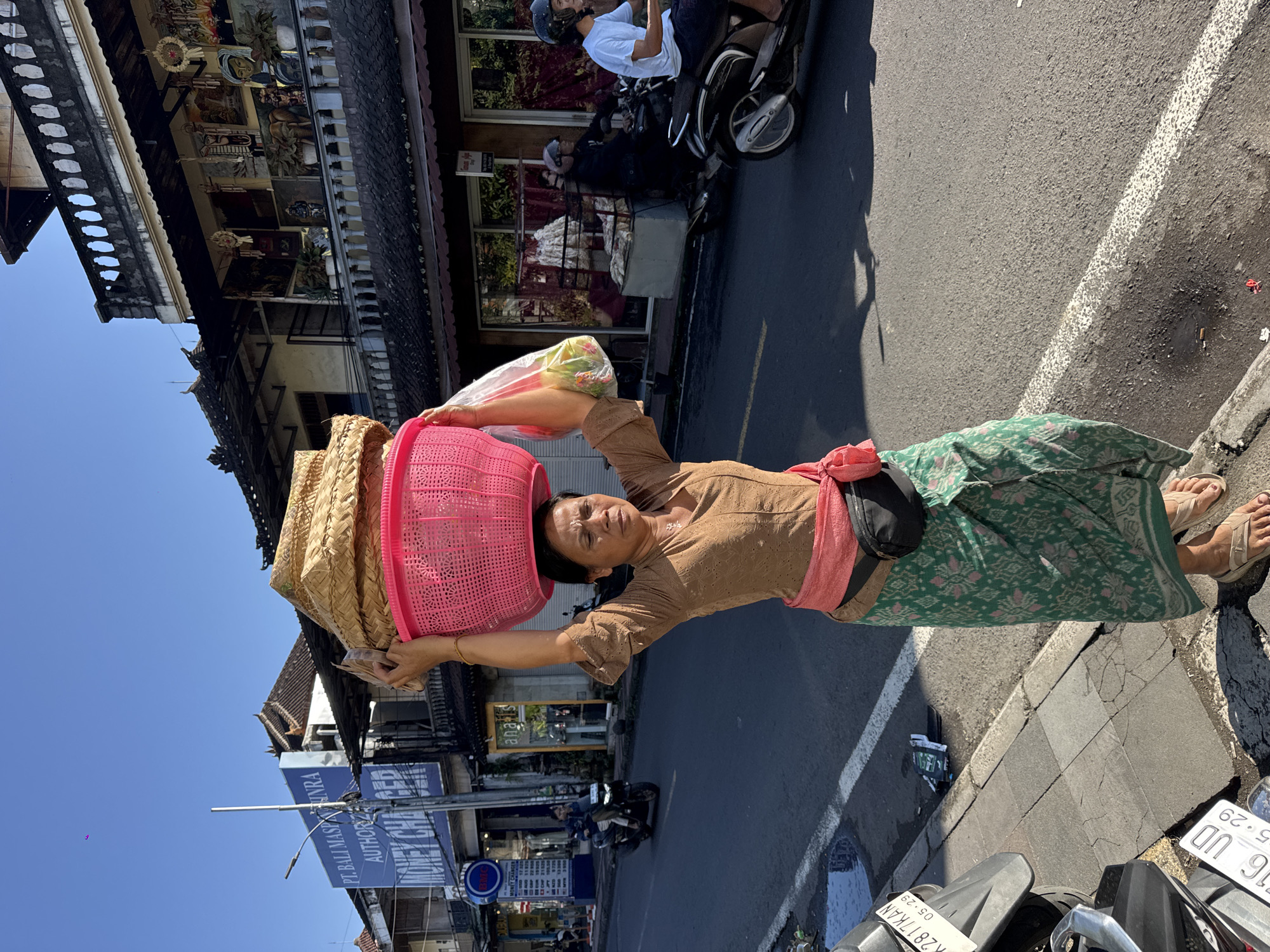 An Indonesian woman walks along a sunny street in Ubud, Indonesia, carrying a pink plastic basket filled with straw items on her head. She wears a brown, long-sleeved, textured blouse, a pink sash tied around her waist, and a green patterned sarong. A black fanny pack is secured around her waist over the sash. She has sandals on her feet and a yellow bracelet on her left wrist. In her right hand, she holds a clear plastic bag with items inside. The street has a paved road with white lines and a sidewalk. Other people and vehicles, including motorbikes, are visible in the background. Buildings with traditional architecture line the street. One building has a sign that reads "PT. BALI MASPURA AUTHORIZED MONEY CHANGER".