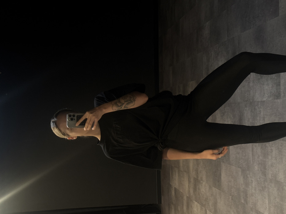 An individual is depicted indoors, capturing a mirror selfie with a light-colored smartphone that obscures their face. The subject has light-colored hair and a floral tattoo on their left forearm. They are wearing an oversized black t-shirt, tied in a knot at the front, and tight black leggings. A ring is visible on the fourth finger of their right hand. The background consists of a plain, dark-colored wall. The floor is composed of large, rectangular, gray-toned panels arranged in a staggered pattern. The lighting is subdued, with a bright reflection of an overhead light source visible in the upper left portion of the mirror. The environment presents as a minimalist indoor space, such as a fitness studio or dance room. No specific visual cues within the image definitively identify the location as Dubai, United Arab Emirates.
