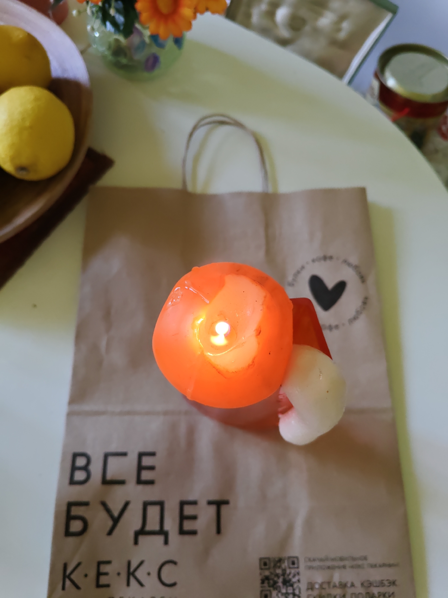 A close-up, top-down view captures a table setting with a brown paper bag, a lit orange candle, and a white and red mushroom-shaped object resting on it. The bag displays text and a black heart emblem. To the left, a wooden bowl holds two bright yellow lemons. In the background, a vase with orange flowers and a jar with a red lid are visible. The scene appears to be set indoors. There is no discernible information indicating a specific location within the city of Rostov-on-Don, Russia.