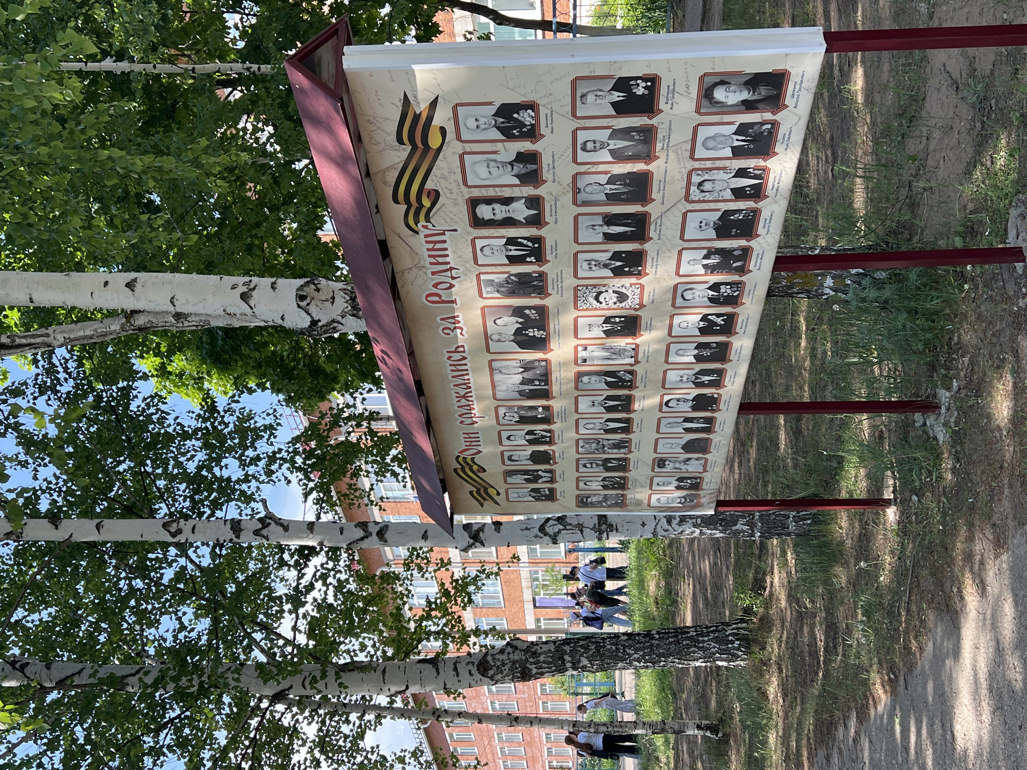 The image depicts a memorial display board in a park-like setting with birch trees. The board features a collection of black-and-white portraits of people, possibly veterans or war heroes, organized in a grid. A ribbon is visible on the top left of the board. The text on the board is in Russian and reads "Они сражались за Родину", translating to "They fought for the Motherland". In the background, there is a brick building, and several people are walking. The location is Noviy, Russia.