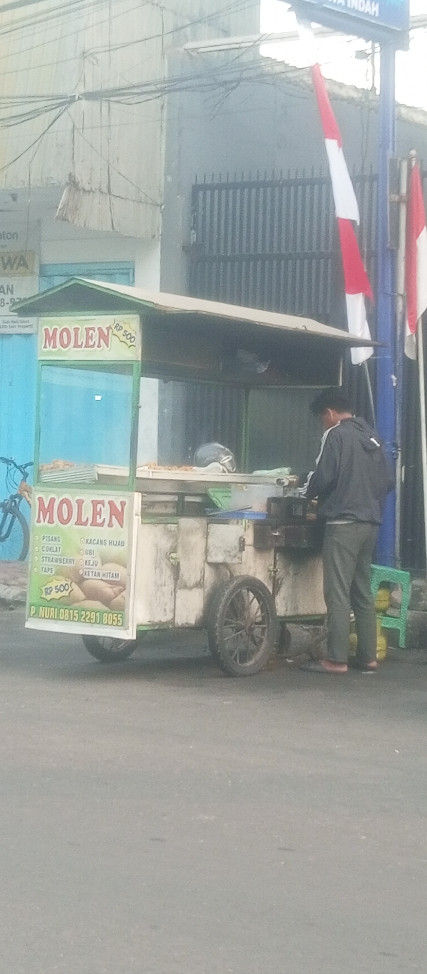 The image depicts a street scene in the city of Kediri, Indonesia, likely in an urban or commercial area, given the presence of a food cart and adjacent buildings. The primary subject is a food cart selling "Molen," a type of fried dough snack, with a visible price of RP 500. The cart is white with green accents and has a transparent display showcasing the products, along with a menu listing various fillings such as "PISANG" (banana), "COKLAT" (chocolate), "STRAWBERRY", "TAPE" (cassava tape), "KACANG HIJAU" (mung bean), "UBI" (sweet potato), "KEJU" (cheese), and "KETAN HITAM" (black sticky rice). A person, likely the vendor, is seen operating the cart from the side, wearing a dark hooded jacket and gray pants. The background shows a building with a corrugated metal facade and some electrical wiring, as well as Indonesian flags. A bicycle is parked to the left of the food cart, and a green plastic stool is visible on the right. The road surface appears to be asphalt. The overall impression is a typical street vendor scene in Indonesia.
