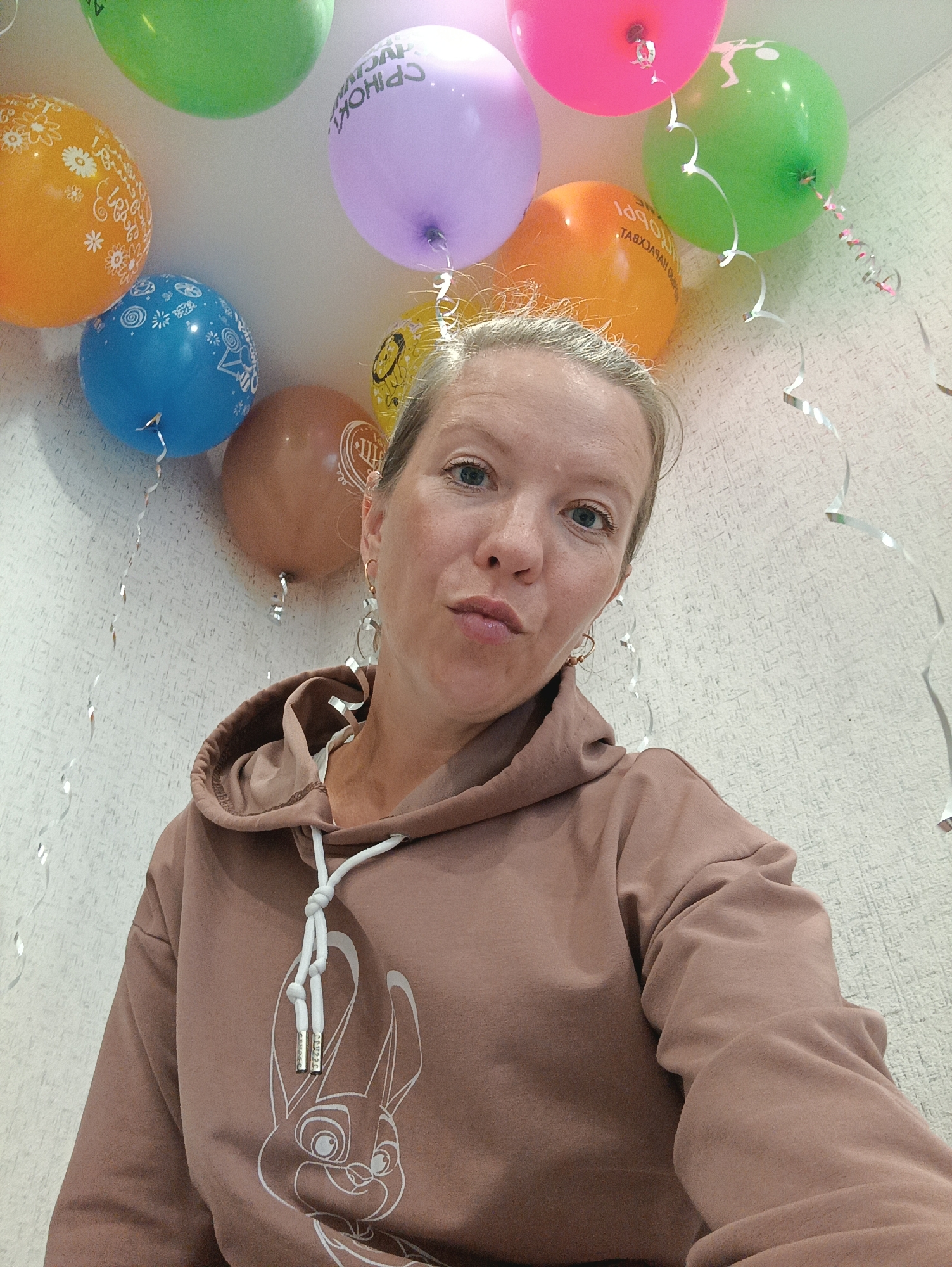 An adult female is depicted in a close-up selfie, captured indoors in what appears to be a residential setting within the city of Chaikovsky, Russia. She has light-colored hair pulled back, light-colored eyes, and a neutral expression with slightly puckered lips. She wears a brown hooded sweatshirt featuring a white line-art graphic of a rabbit's face on the chest and white drawstrings with metallic aglets. The background consists of a light-colored, textured wall adorned with numerous colorful balloons and silver curling streamers. Visible balloons include green, light purple, pink, orange, blue, brown, and yellow. The light purple balloon clearly displays the Russian text "С ДНЕМ РОЖДЕНИЯ" (Happy Birthday). Other balloons feature white decorative patterns or text. The overall scene suggests a birthday celebration.