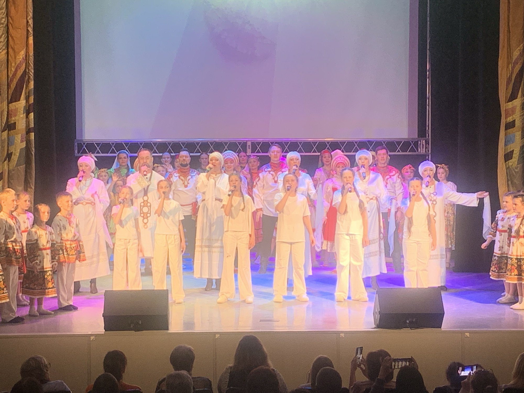 A group of people, likely a choir or performance group, are standing on a stage in what appears to be the city of Chaikovsky, Russia. The stage is set with a large screen backdrop and speakers at the front. The people are dressed in a mix of traditional Russian clothing and modern white outfits. Some are holding microphones and appear to be singing. There are children and adults present, suggesting it could be a multi-generational group. The audience is visible in the foreground, with some people holding up phones, likely to take photos or videos of the performance. There are curtains on the side of the stage. The lighting suggests an indoor venue, possibly a theater.