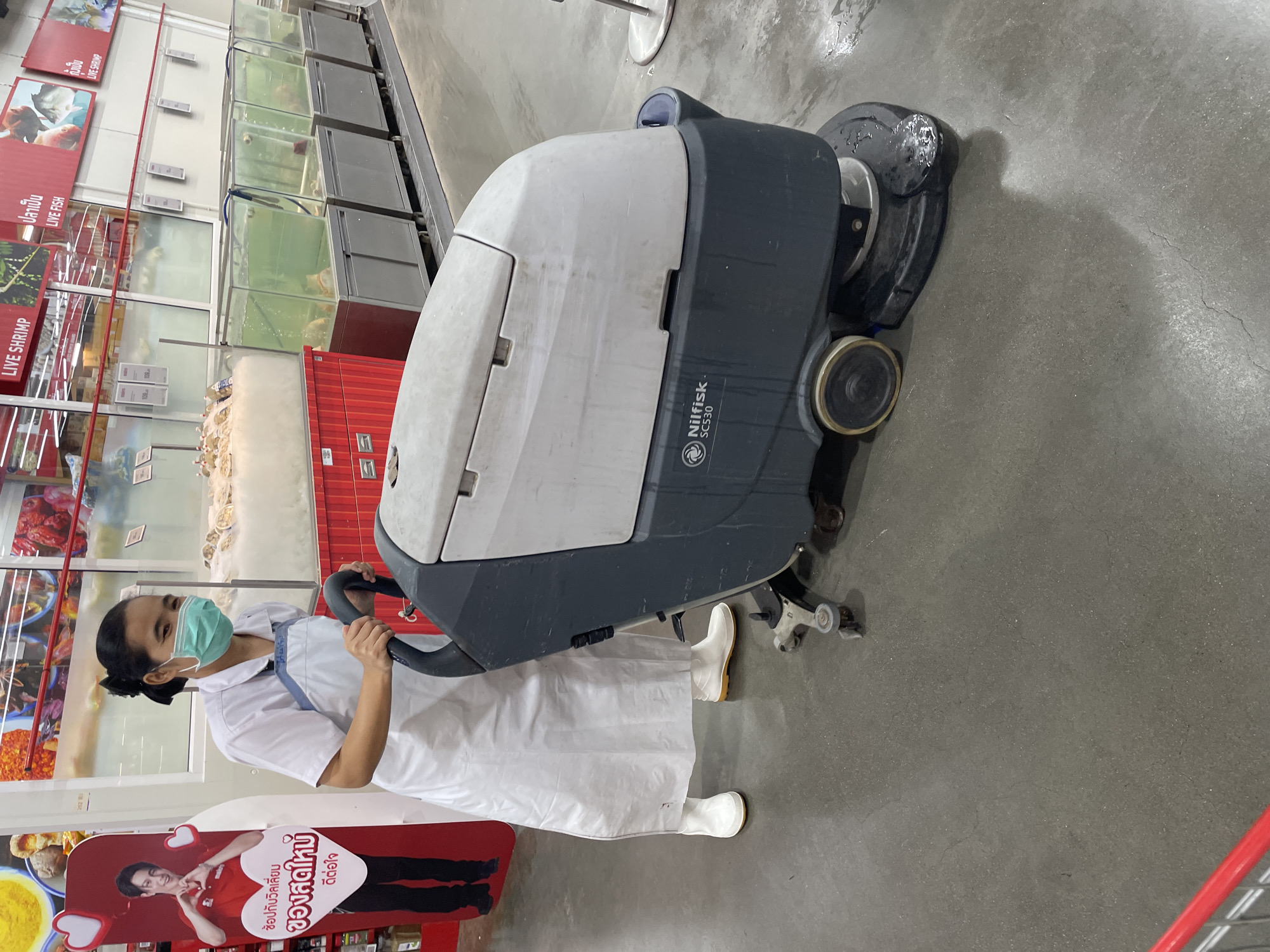 In Pattaya City, Thailand, a female employee wearing a white uniform, blue apron, and a green face mask is operating a Nilfisk SC530 floor cleaning machine. She is holding the machine's handle and appears to be pushing it forward. The environment is an indoor commercial space, likely a supermarket or large retail store, with various food displays visible in the background. These include a refrigerated seafood display with tanks of live fish and a display stand featuring a person making a heart shape with their hands. The floor is concrete and appears wet in some areas, suggesting the cleaning activity is in progress. The overall scene depicts a maintenance activity within a commercial setting.