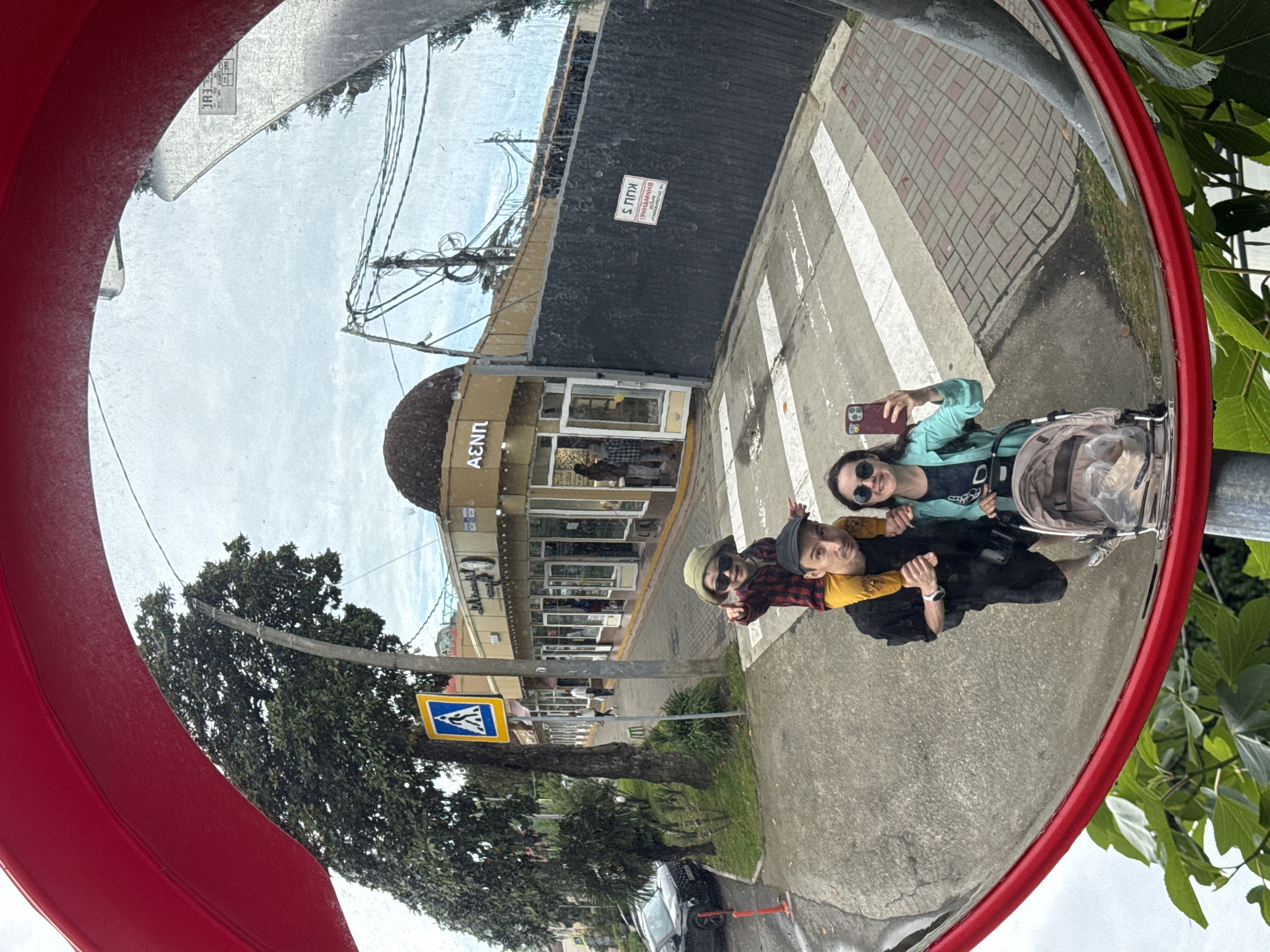A fisheye lens perspective captures a street scene via a red, circular traffic mirror. Three people—two women and one man—stand close to the mirror, visible as reflections. The woman closest to the mirror is holding a smartphone up, possibly taking a picture. They stand on a sidewalk next to a crosswalk, indicated by white stripes painted on the road. Behind the group are buildings with storefronts. Above the buildings, power lines stretch against a light, cloudy sky. A road sign indicating a pedestrian crossing is visible on the left. Vegetation is scattered throughout, including a large tree on the left and foliage around the mirror on the right. The location is Sochi, Russia, as a sign on a building in the background is written in the Cyrillic alphabet.