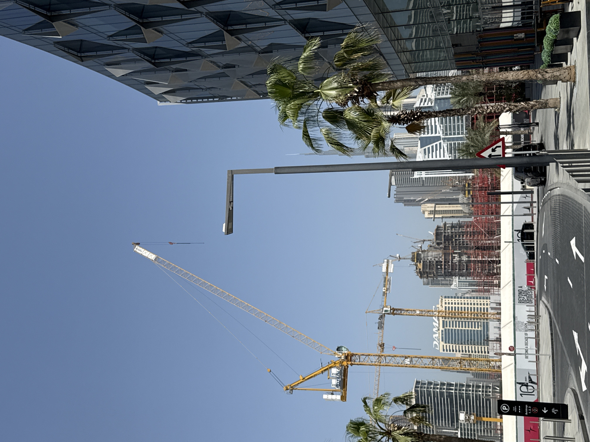The image depicts a scene in Dubai, United Arab Emirates, characterized by rapid urban development and construction. In the foreground, there is a street with palm trees lining the sidewalk. Tall modern buildings flank the street, with a prominent building featuring a geometric, pyramid-like pattern on its facade.
Several construction cranes are visible in the distance, indicating ongoing construction projects. One particularly large crane dominates the left side of the frame against a clear blue sky. The skyline is filled with a mix of completed buildings and structures under construction, showcasing Dubai's architectural landscape.
The street appears to be modern, with road markings and signage visible. The overall impression is one of a bustling urban environment with a focus on growth and modernity.