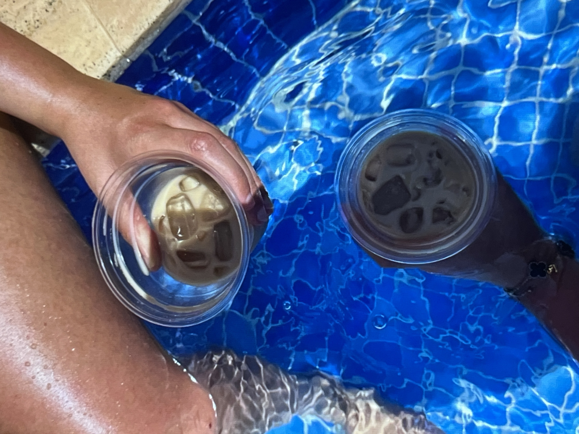 The image shows two hands holding plastic cups filled with iced coffee. The cups are submerged in a clear blue body of water, likely a swimming pool, with a tiled bottom. The water is illuminated from above, casting light and reflections on the surface. One hand is in the foreground, showing smooth skin and a light-colored bracelet with a clover-shaped charm. The other hand, partially visible on the right, is darker and appears to be holding the second cup. The setting suggests a relaxing or leisurely moment, possibly a vacation or a warm day. The lighting indicates daytime.