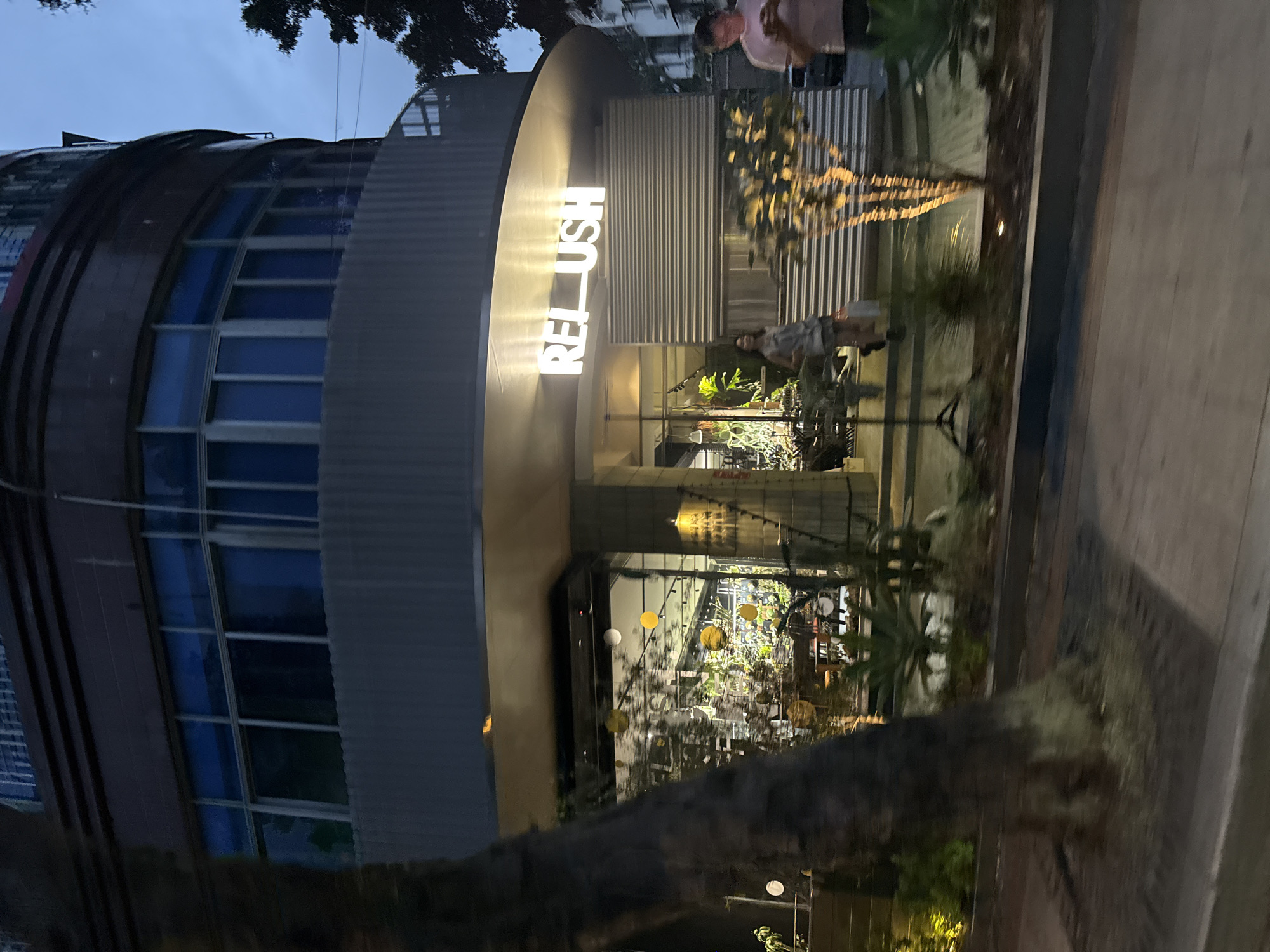 The image captures a nighttime street scene in Haizhu District, China, featuring a building with a distinctive cylindrical shape and a curved facade. The building is illuminated by the glowing "REL_USH" signage above the entrance. A person in a pink shirt stands near the right side of the building, while another individual is visible closer to the building's entrance. The building's windows reveal interior lighting and suggest the presence of decorative elements. The foreground includes foliage and a tree trunk, adding a natural element to the urban scene. A paved sidewalk is also visible.