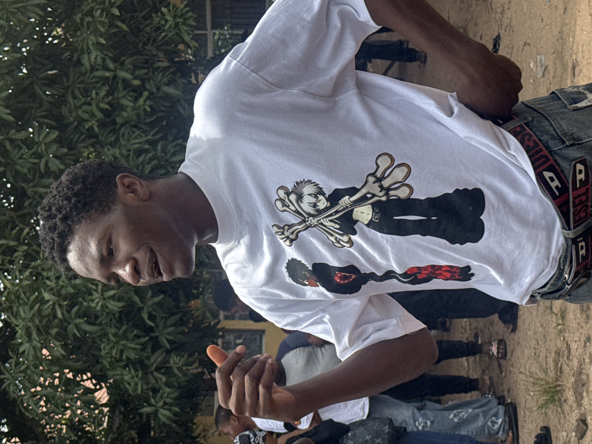 In Ihiagwa, Nigeria, a young Black male stands outdoors, facing right and speaking. He is wearing a white t-shirt with a graphic print of two anime-style characters, one with black hair and a red-and-black patterned garment, and the other with white hair holding a bone-like object. He also wears a denim belt with a red "Diesel" logo buckle. His right hand is raised, with his index finger extended, as if emphasizing a point. Behind him, other people are visible, some blurred, and lush green foliage suggests a natural or garden setting. The ground appears to be dirt and sand.