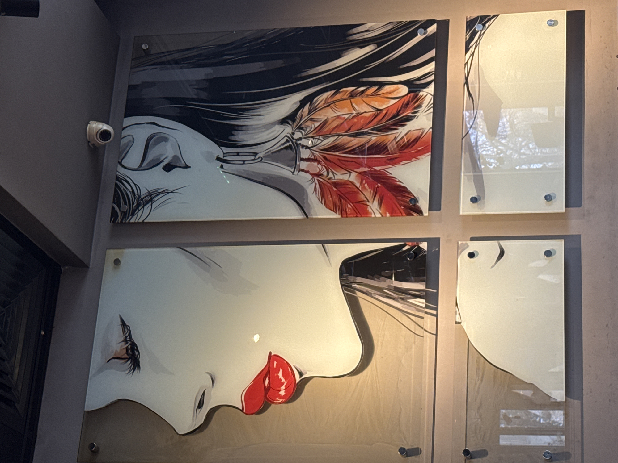 Four panels are arranged on a gray wall, each attached with metal studs. The top left panel depicts a stylized drawing of a person's face in profile, with dark hair and orange and red feathers incorporated into the hair design. The bottom left panel shows another stylized drawing of a face in profile with red lips. The top right and bottom right panels have a curved, reflective design, making them look like mirrors, reflecting the outside environment. A surveillance camera is visible on the wall above the top left panel. The scene is located in Sofia, Bulgaria.