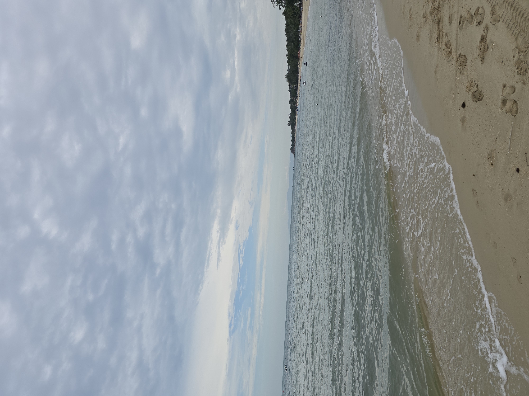 A wide shot of a sandy beach with calm, grey-green water lapping at the shore under a cloudy sky. The sand in the foreground is wet from the receding tide and shows several footprints. The water stretches out to the horizon where a line of green trees and buildings is visible. In the distance, there are a few small boats and people dotting the shoreline and water. The overall impression is a quiet, overcast day at the beach.