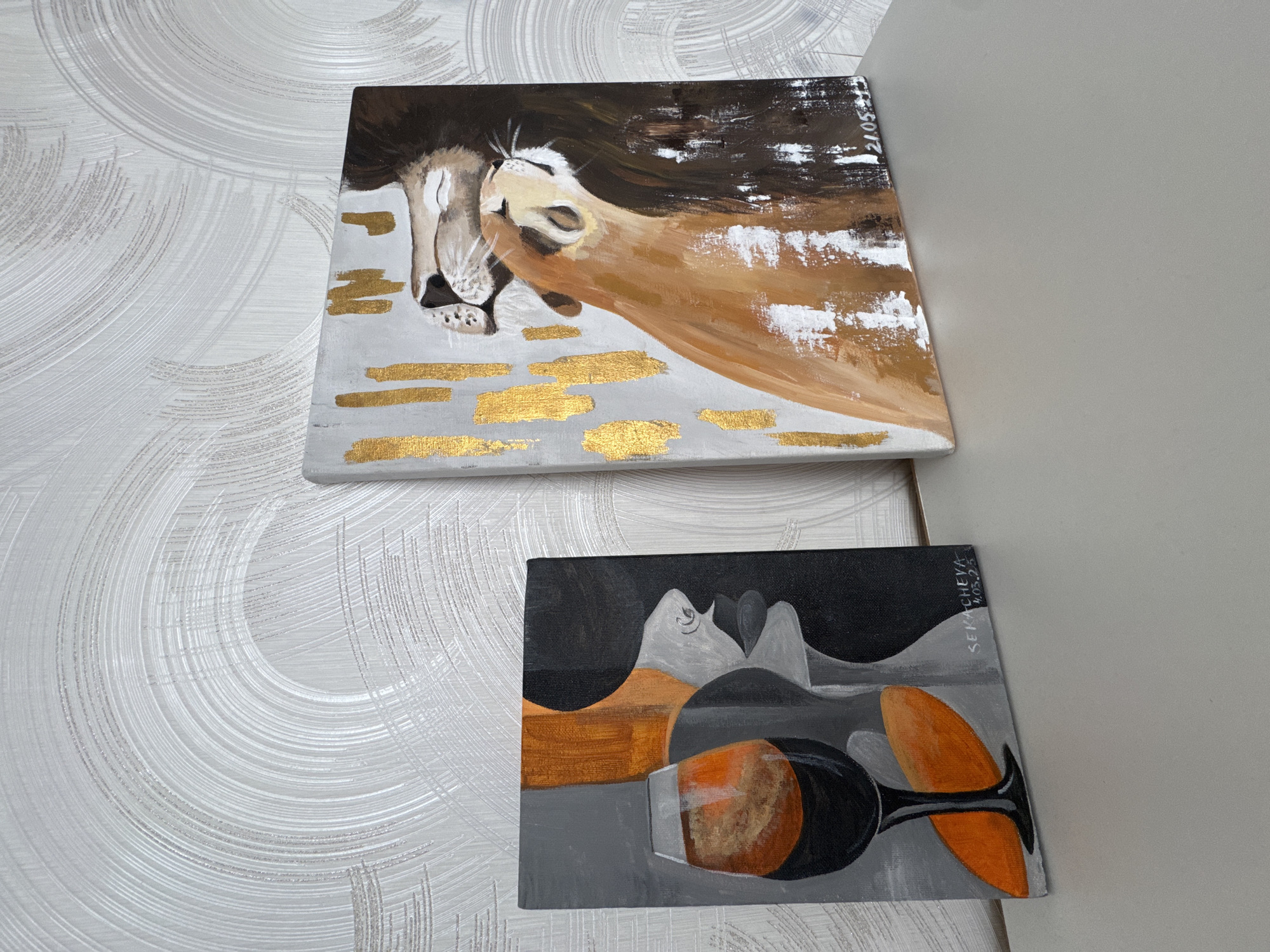 Two paintings are displayed against a patterned white wall, with a section of a light gray wall visible on the right side.
The upper painting depicts two lions in a close embrace, rendered in shades of brown, white, and gold. The canvas appears to have been painted with thick brushstrokes. The date "21.05.2023" is inscribed in the upper right corner of the canvas.
The lower painting, also on canvas, features an abstract composition in gray, black, orange, and gold hues. It appears to depict a wine glass and perhaps other obscured shapes. The name "SERKACHEVA" and the date "4.03.23" are written in the upper right corner.
Given the inscription on the lower painting it might be that the artist is named Serkacheva. The scene appears to be an indoor setting where paintings are displayed, potentially in a home or gallery setting. No information regarding the location in the city of Sochi, Russia is available from the image.