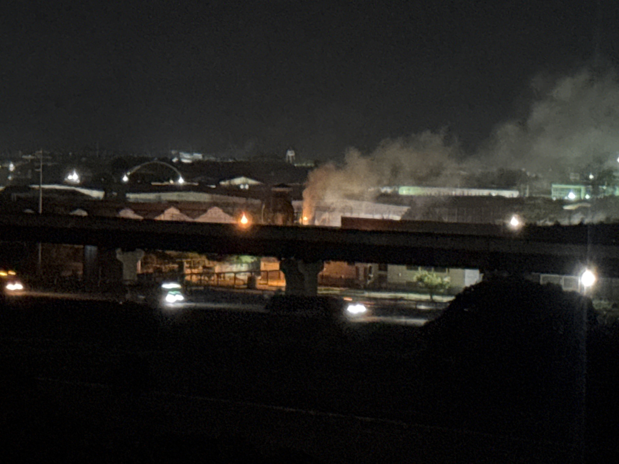 The image shows a dark, urban landscape at night. An elevated bridge spans across the foreground. Car headlights are visible on a road below the bridge. In the background, there is smoke rising from an area with industrial or commercial structures. Lights illuminate the background, suggesting a town or city. Structures like a water tower and a large arch are visible in the distance. Without more information, it is impossible to confirm the location as Maracay, Venezuela.